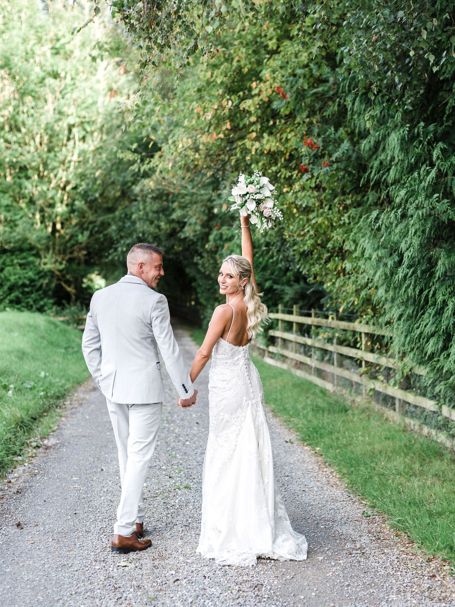 Newlyweds walking hand in hand on country path with bride holding bouquet overhead.