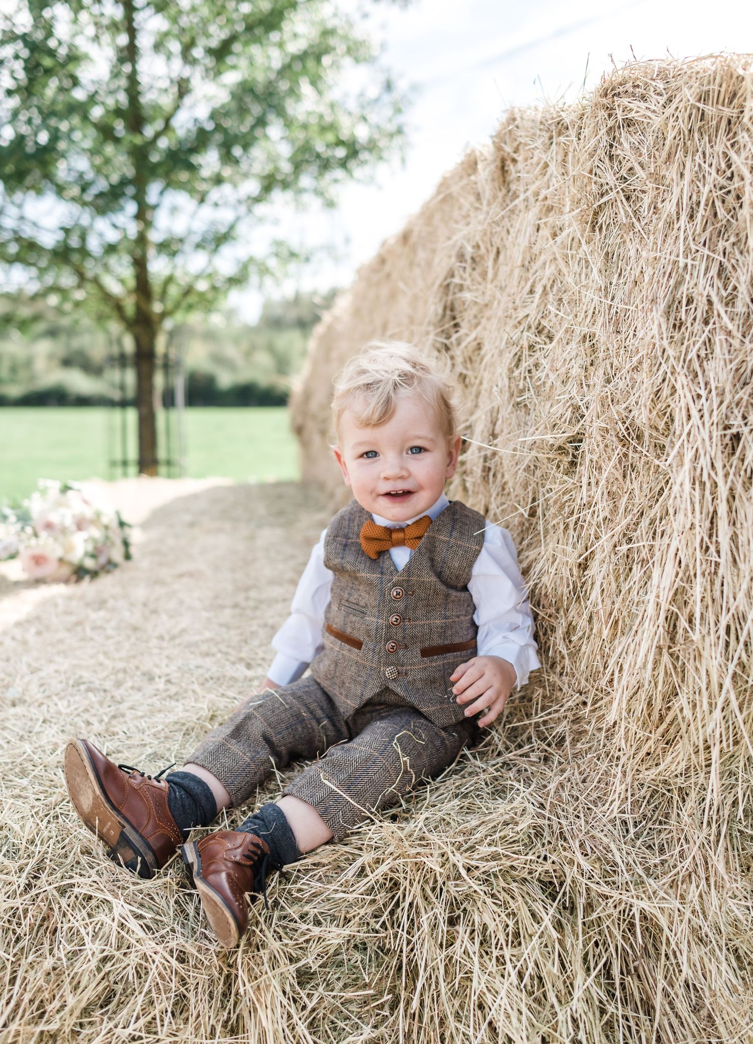 A small child in a tweed vest and bow tie sits on a hay bale in a rustic outdoor setting.