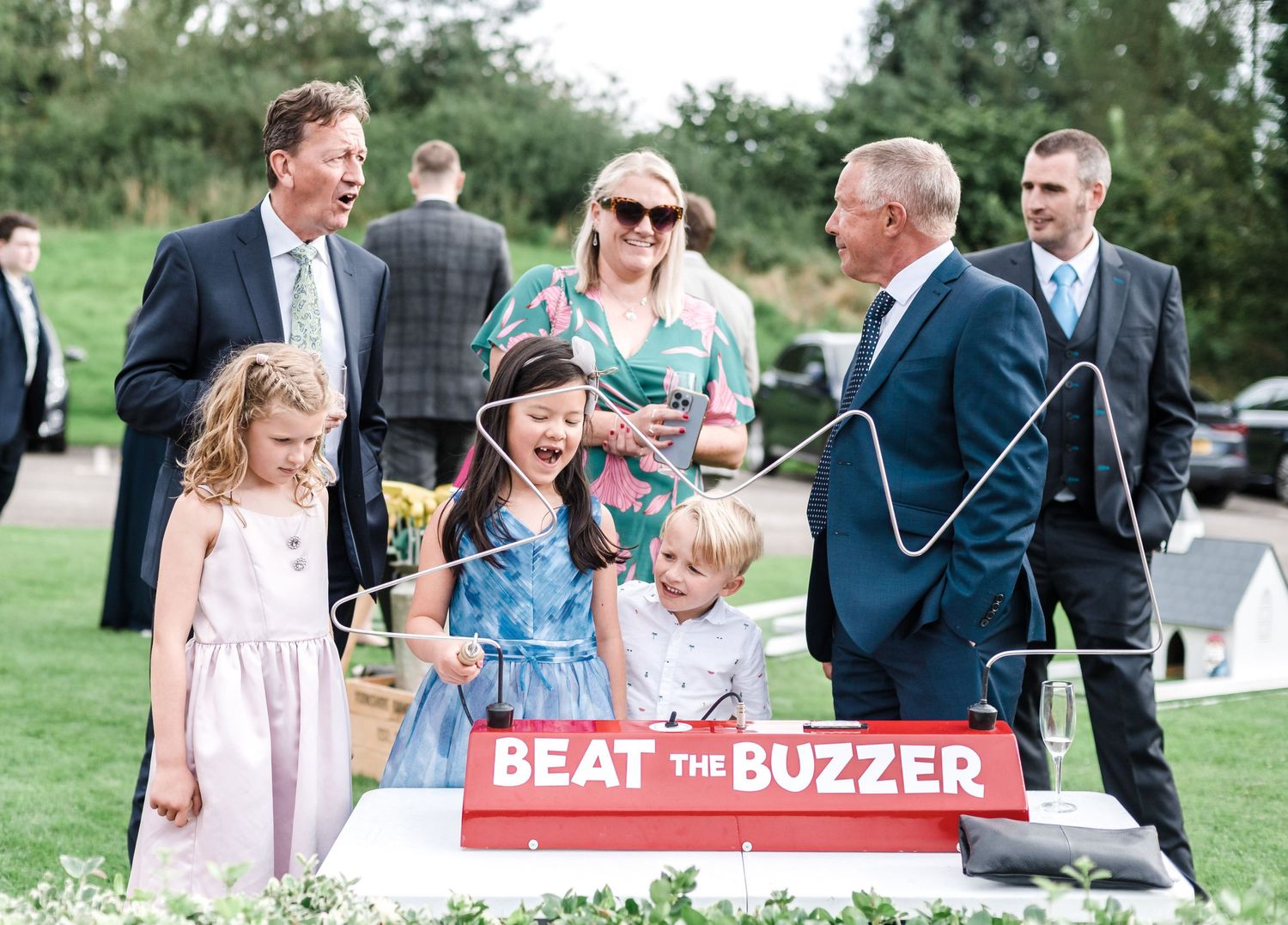 Wedding guests gather around a red 'Beat the Buzzer' game station on a grassy lawn during outdoor reception festivities.