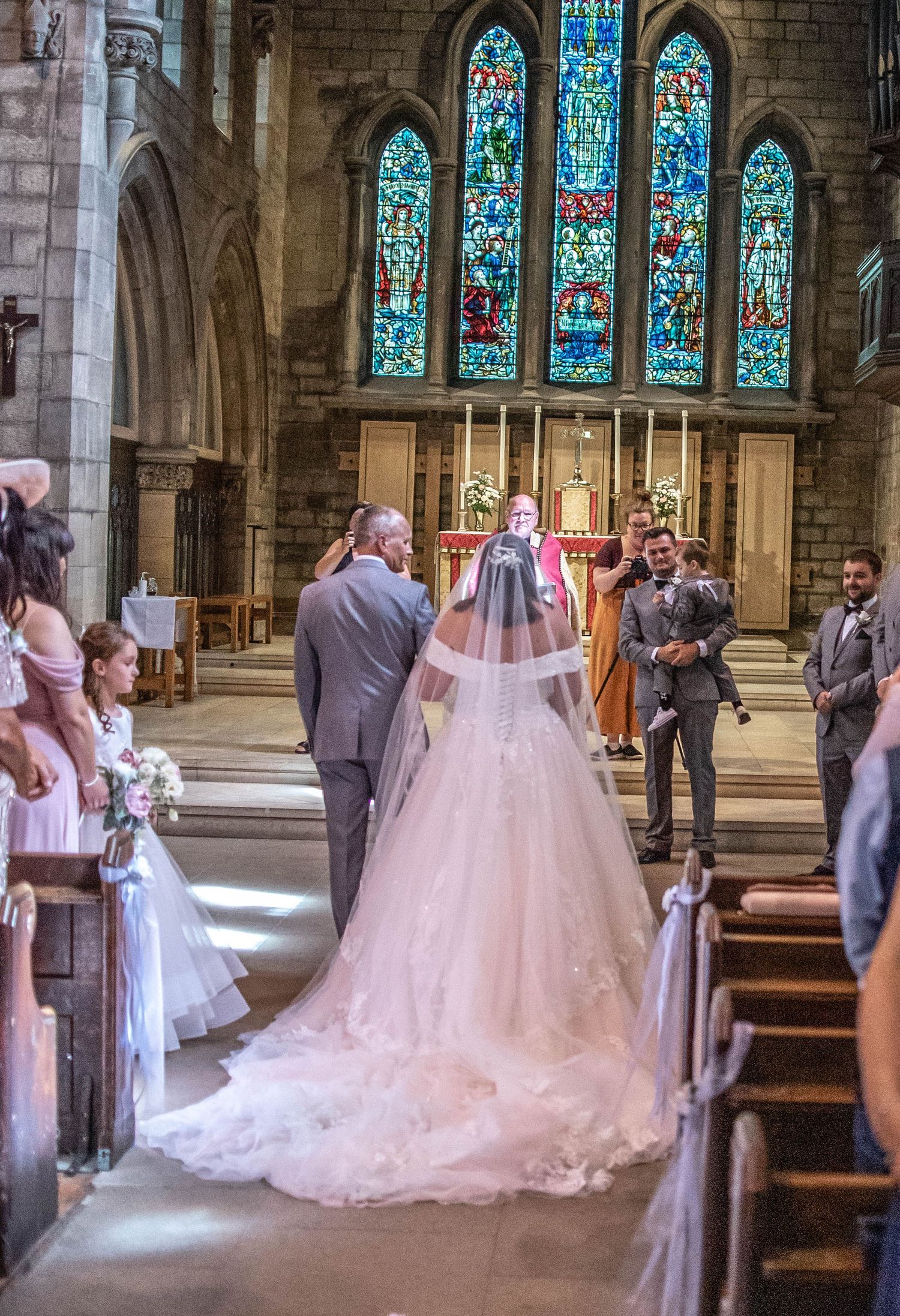 A bride in a flowing gown walks down the aisle of a church with stunning stained glass windows.