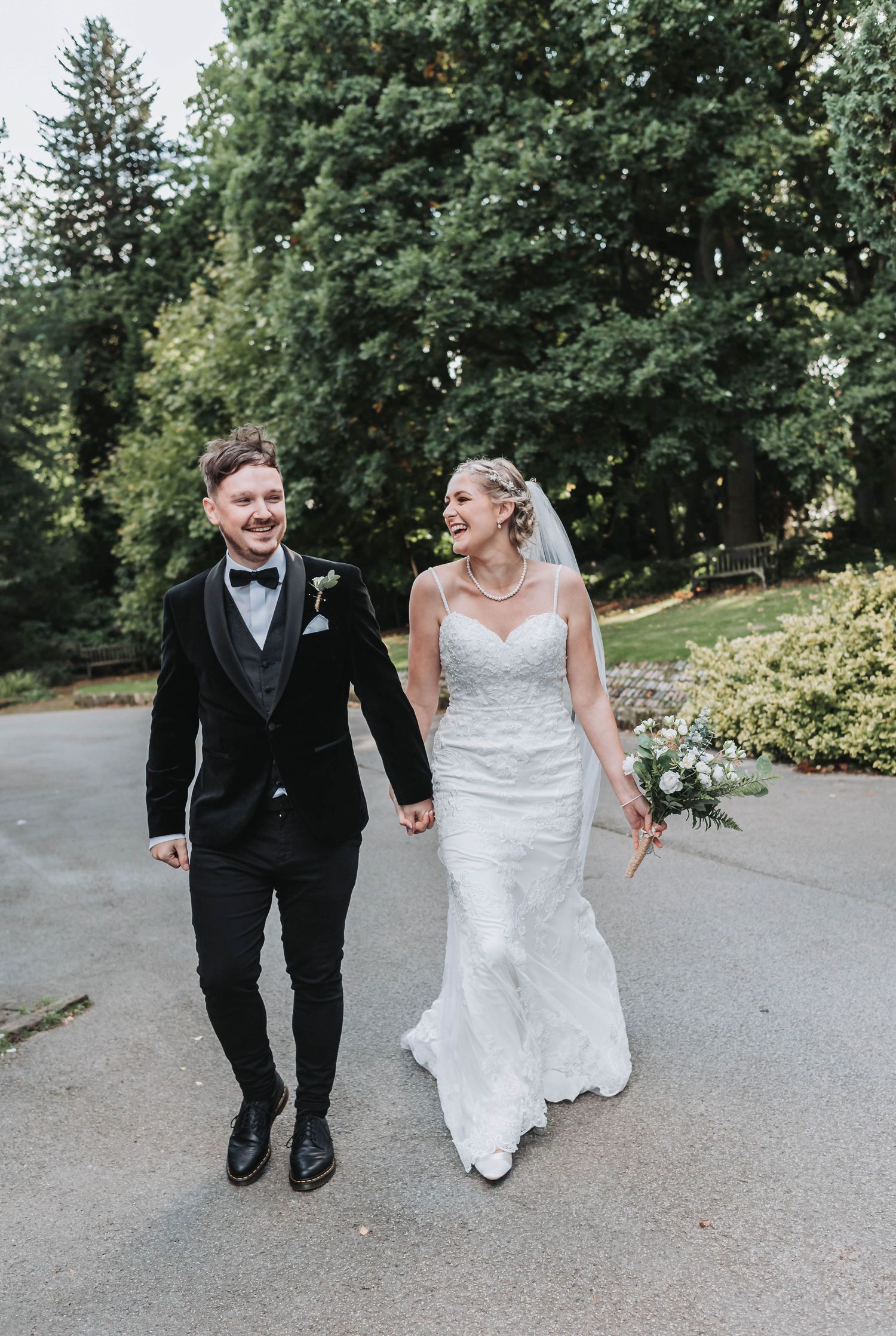 Happy newlyweds walking together on garden path while holding elegant white bouquet.