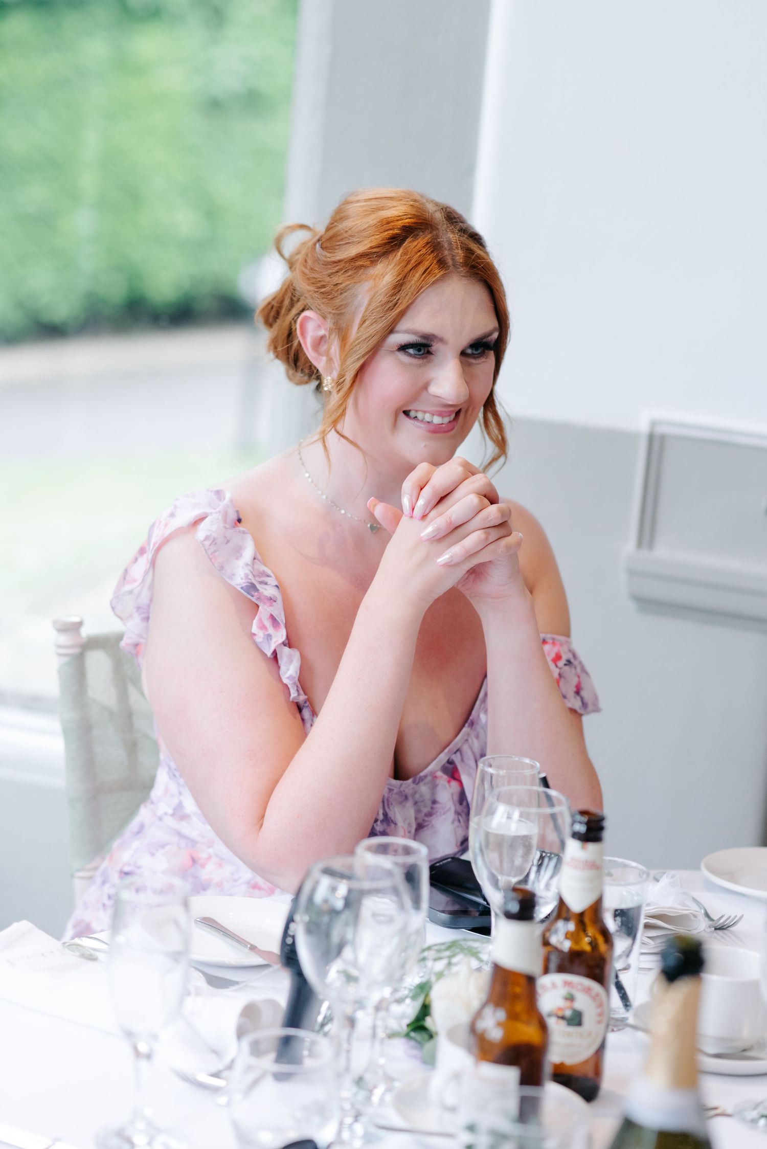 A woman wearing a pink floral dress smiles while seated at a dining table set with glasses and bottles.