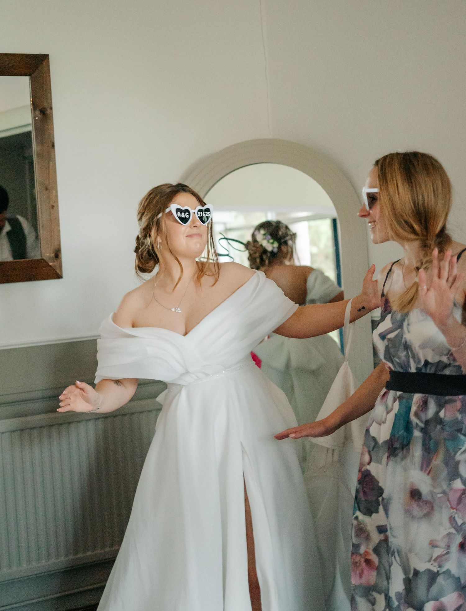 Someone wearing an off-shoulder white wedding dress gets help with buttons and final adjustments during bridal preparation.