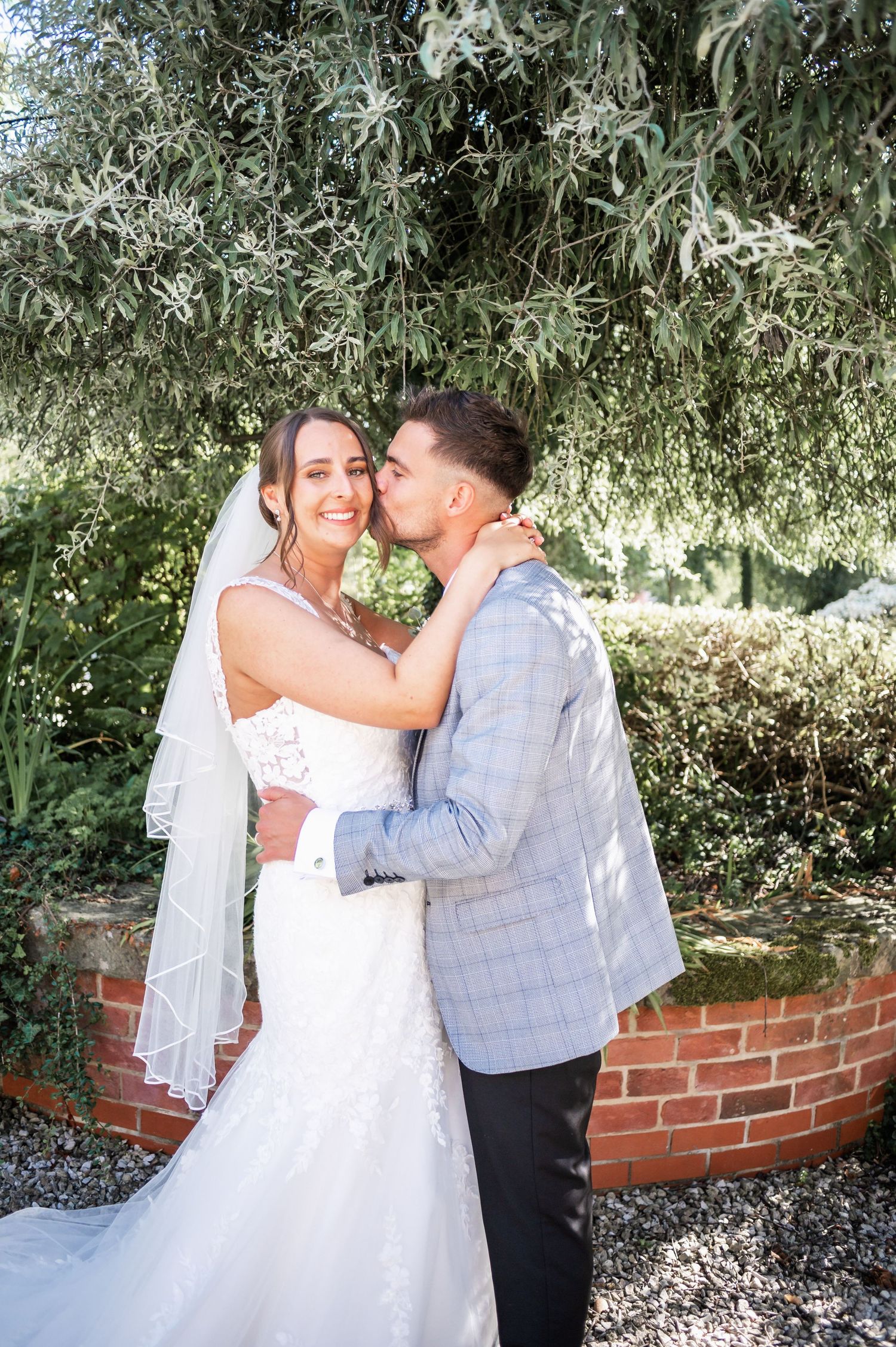 A tender moment between newlyweds beneath willow trees in a garden setting.