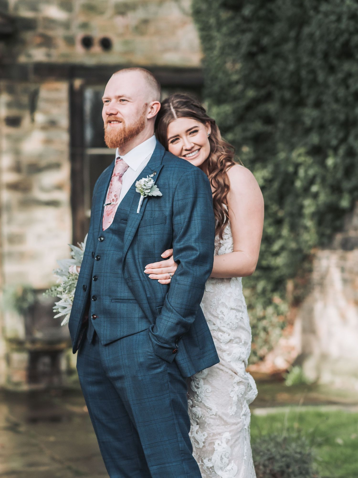 A wedding couple poses together against a stone wall and garden backdrop.