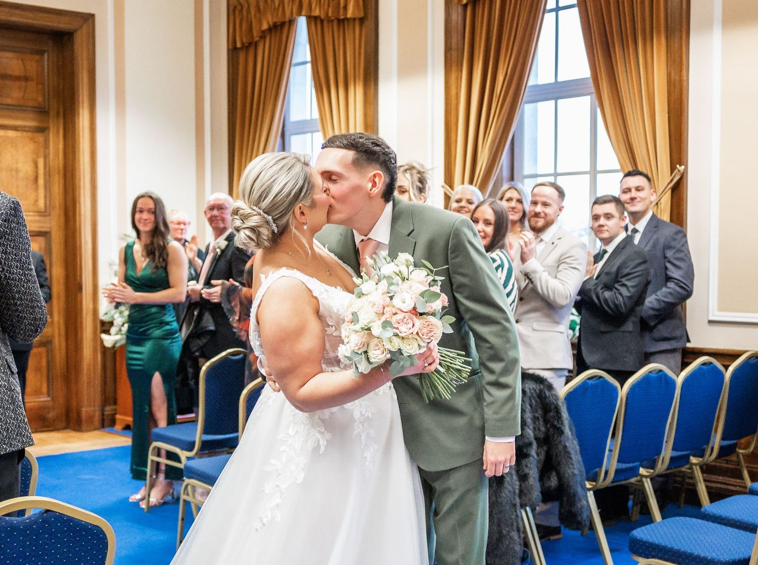 A couple shares their first kiss during a wedding ceremony in an elegant room with gold curtains and blue chairs.