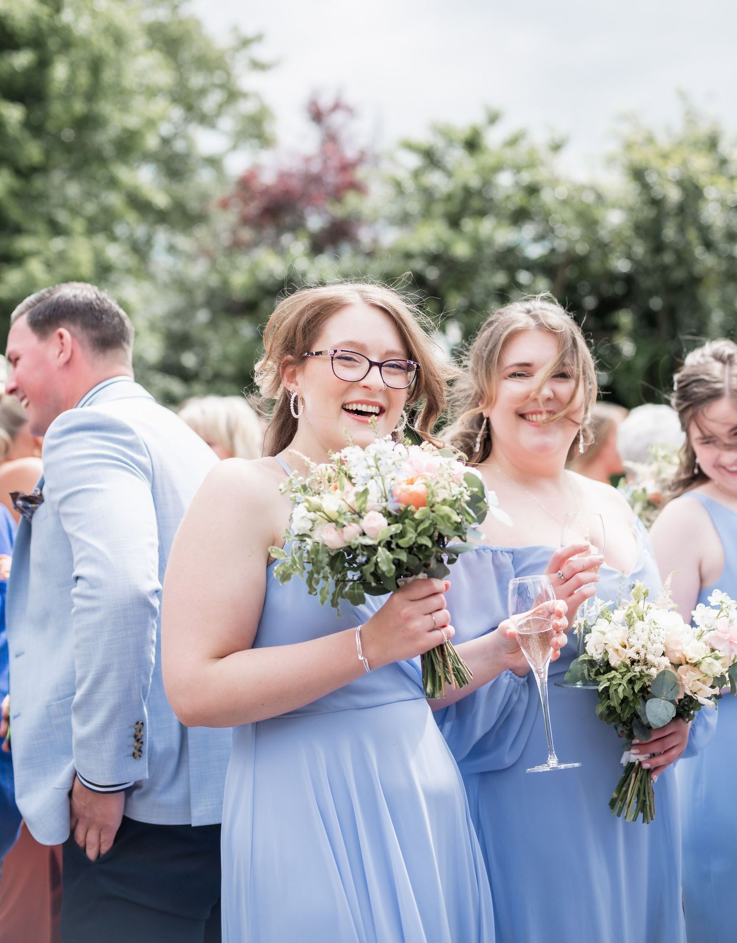 Bridesmaids in powder blue dresses holding bouquets and champagne at outdoor wedding celebration.