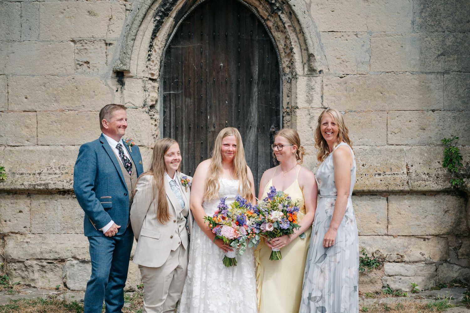 Wedding party poses against stone church wall with rustic wooden door and architectural details.