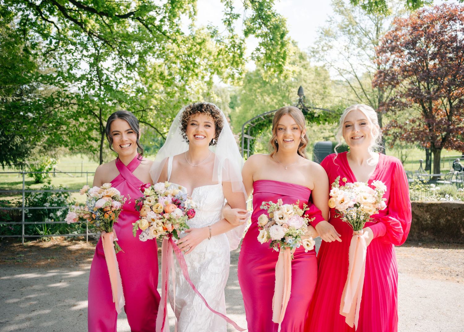 A group in bright pink dresses and white gown stand together holding beautiful floral bouquets in a sunny garden setting.