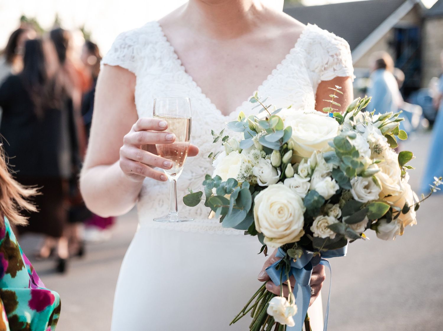 A bride in a white dress holds a champagne glass and elegant white and green floral bouquet during a wedding reception.