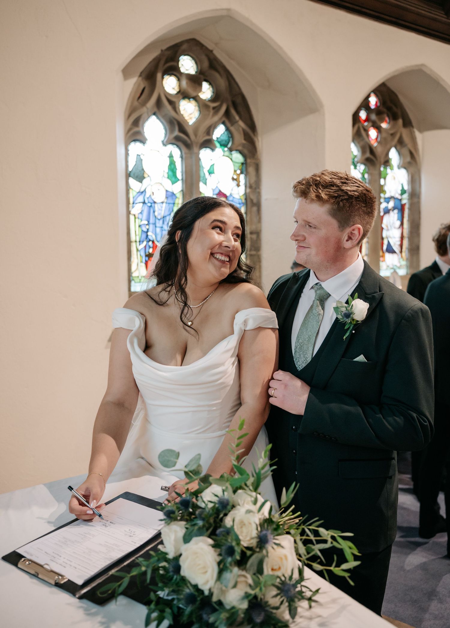Newlyweds sign their marriage certificate during church ceremony with stained glass windows in background.