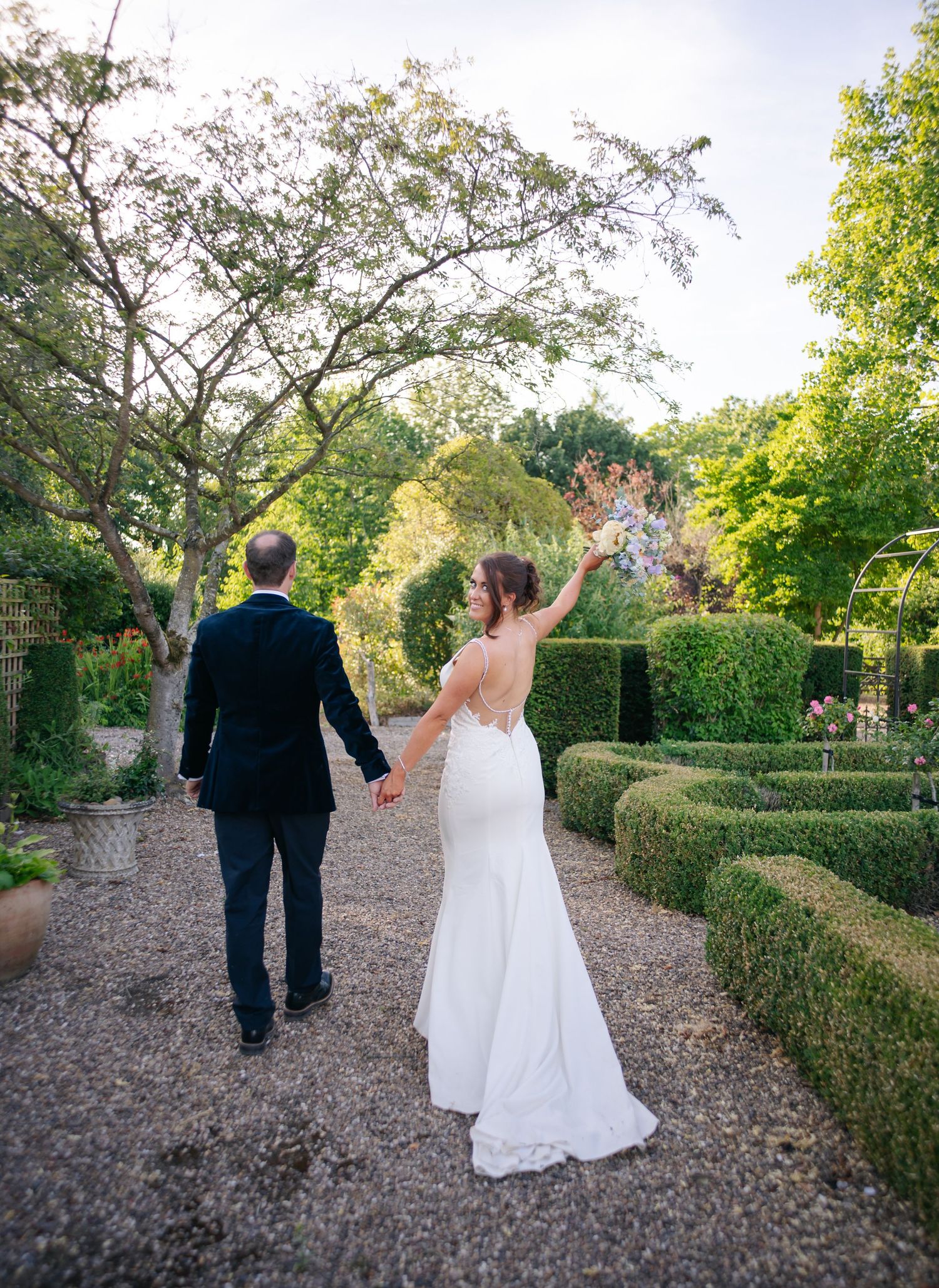 Newlyweds walk hand in hand down gravel path through formal garden with sculpted hedges.