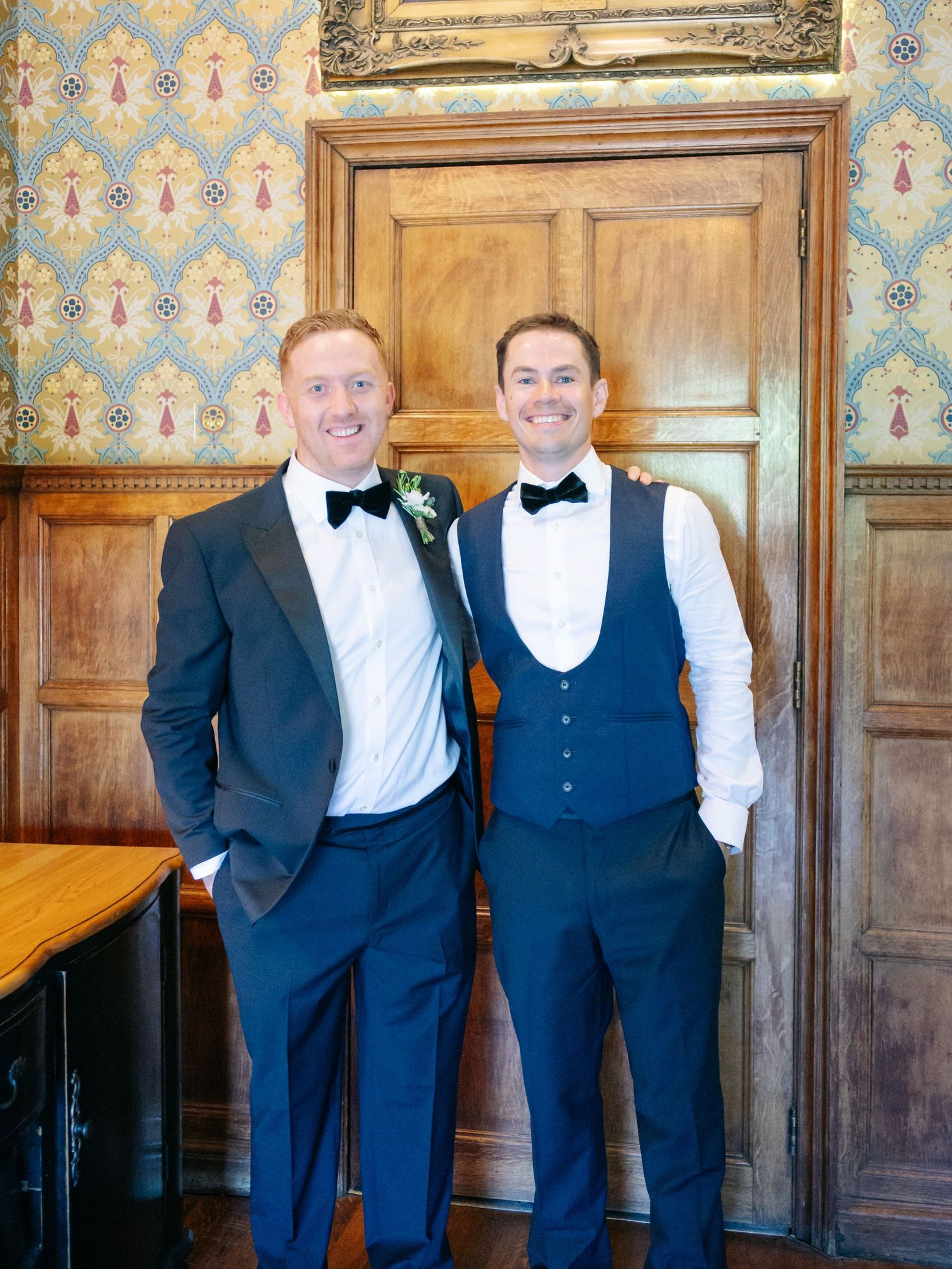 Groomsmen in formal attire posing against wooden paneled wall.