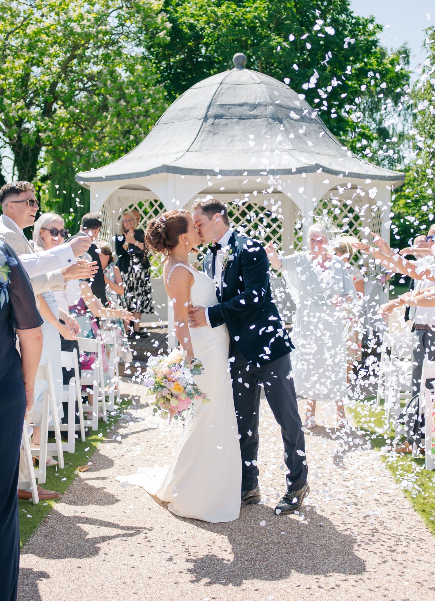 Wedding guests toss white confetti over the newlyweds under decorative garden gazebos.