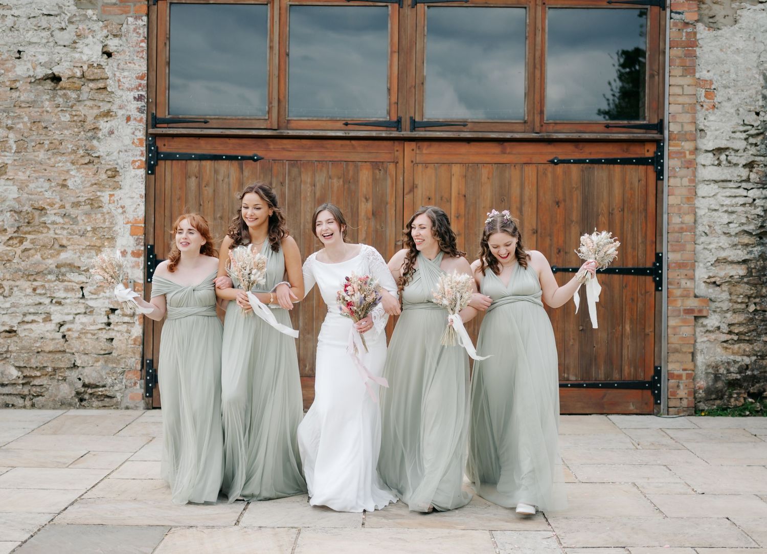 Wedding party in sage green dresses and white gown posing outside rustic brick venue.