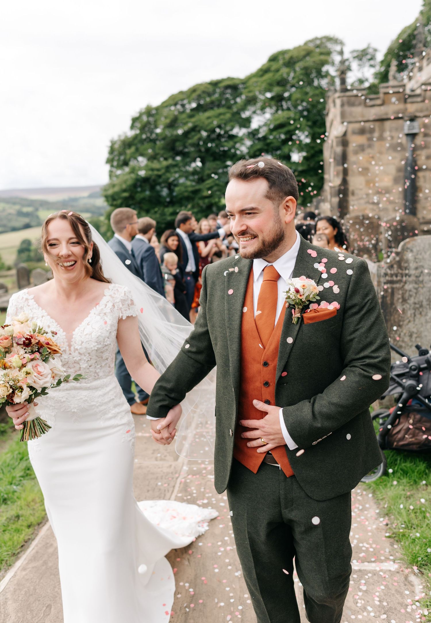 Smiling newlyweds hold hands and walk past cheering guests as confetti falls outside a historic stone church.