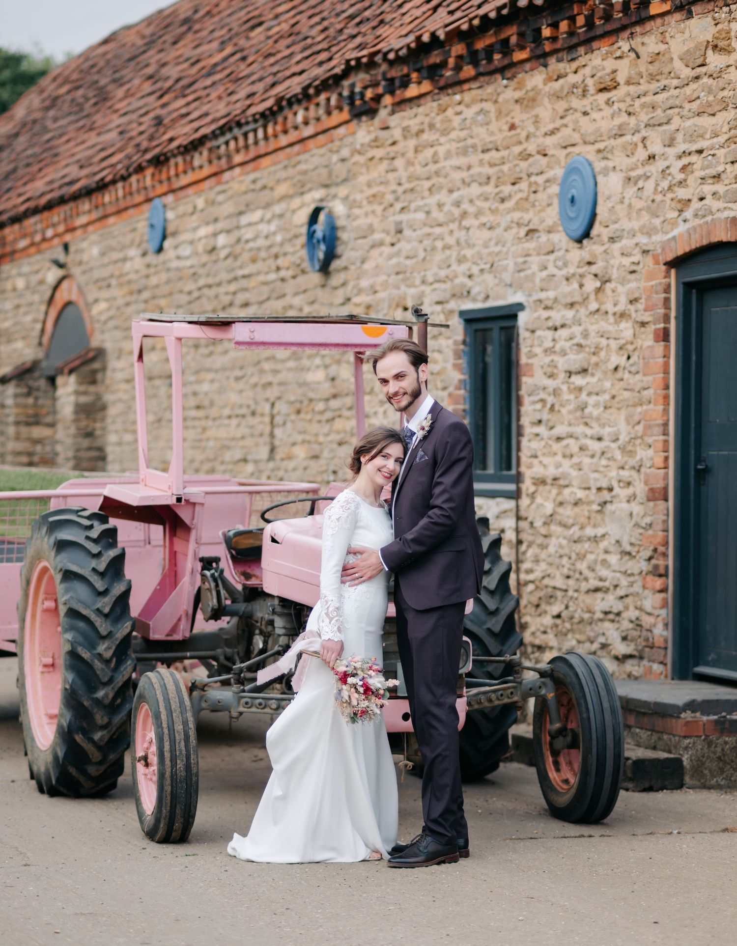 A couple poses in wedding attire next to a vintage red tractor outside a stone barn.