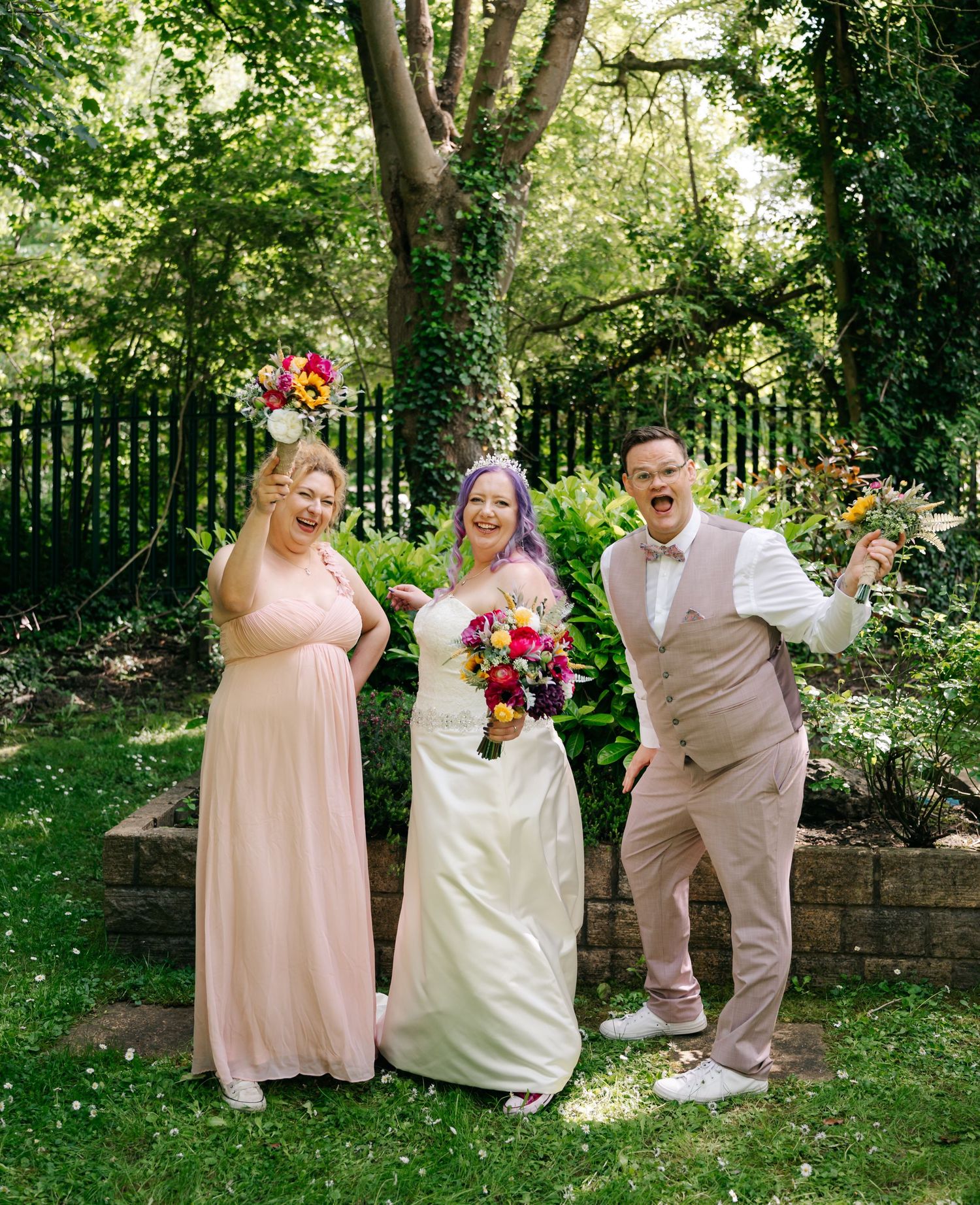 Bride with purple hair holds colorful bouquet, flanked by bridesmaid in pink gown and groom in tan suit outdoors.