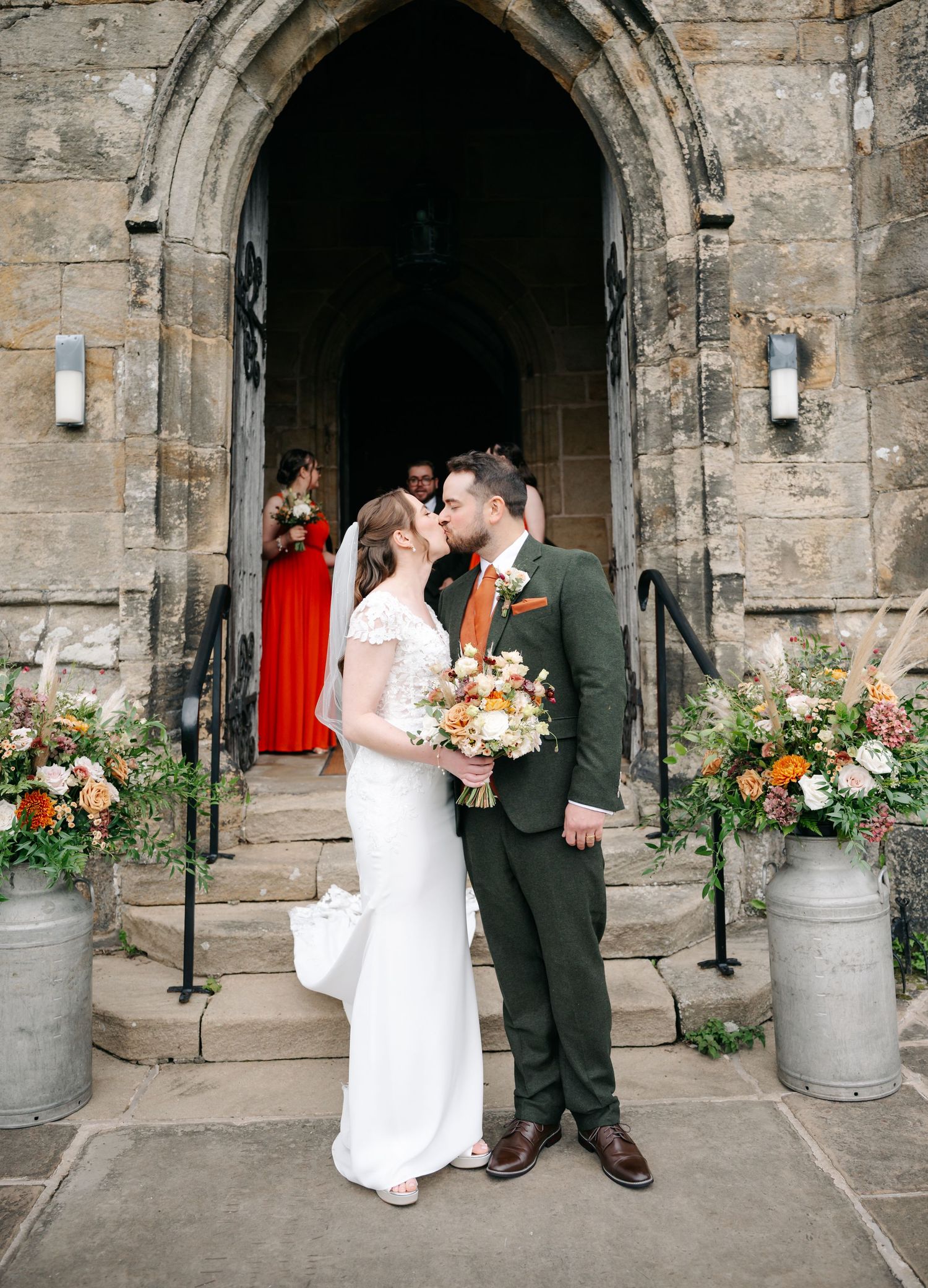 Bride and groom kiss on stone church steps, holding an autumn bouquet, with flower-filled milk churns at the entrance.