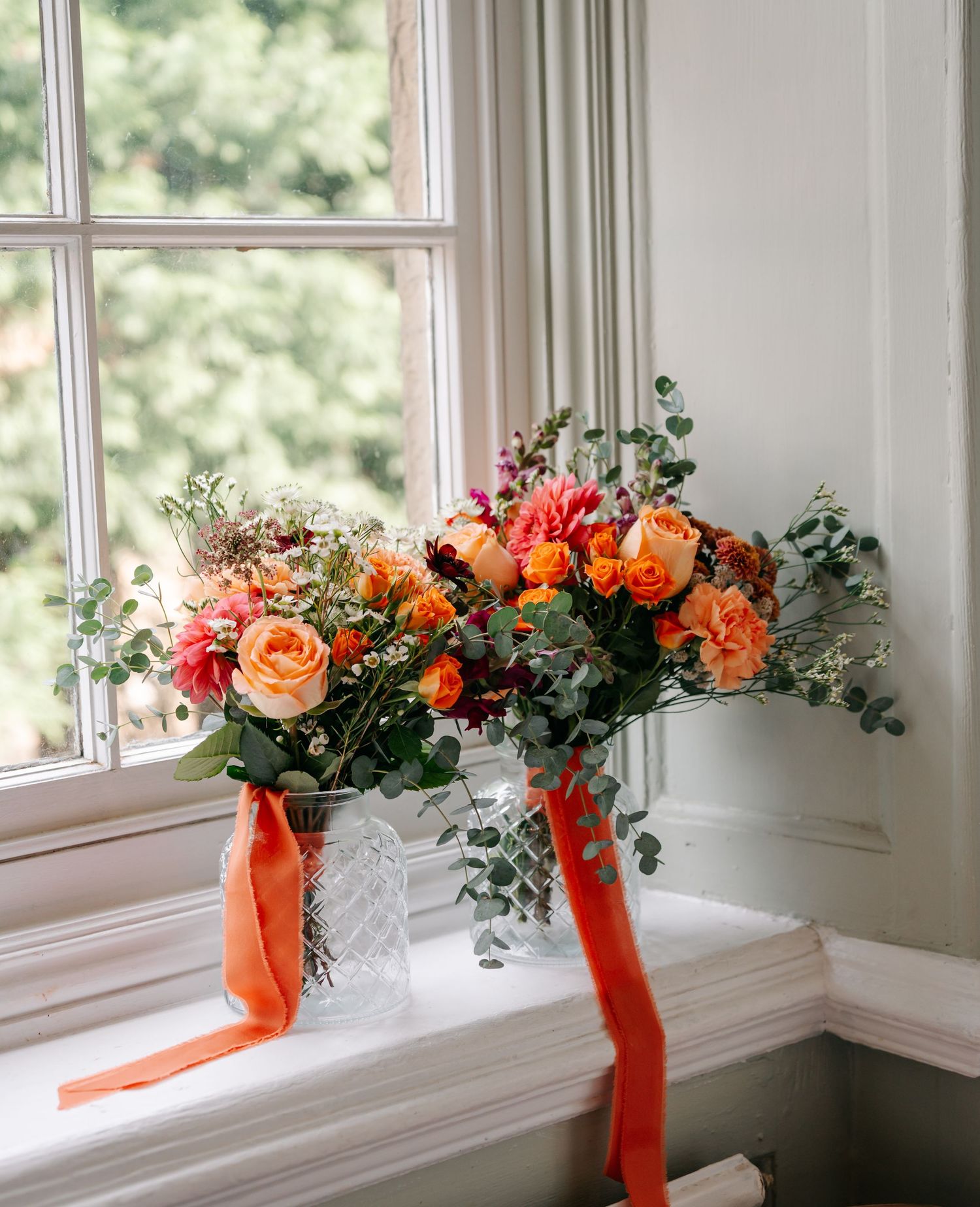 Elegant floral window garland with orange and coral flowers tied with red ribbons decorates white windowsill.