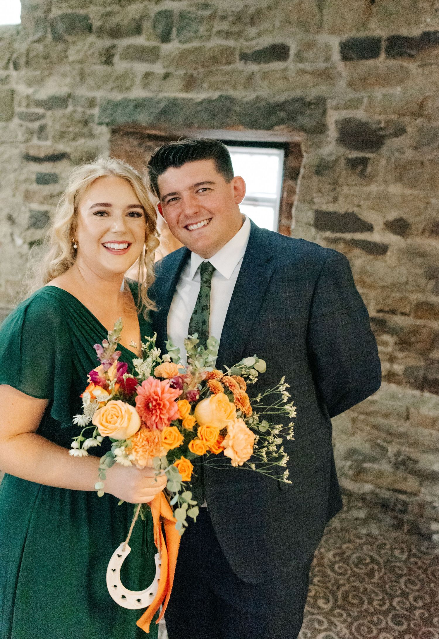 Wedding party members in dark green dresses and navy suits pose with vibrant orange and yellow bouquets against stone wall.
