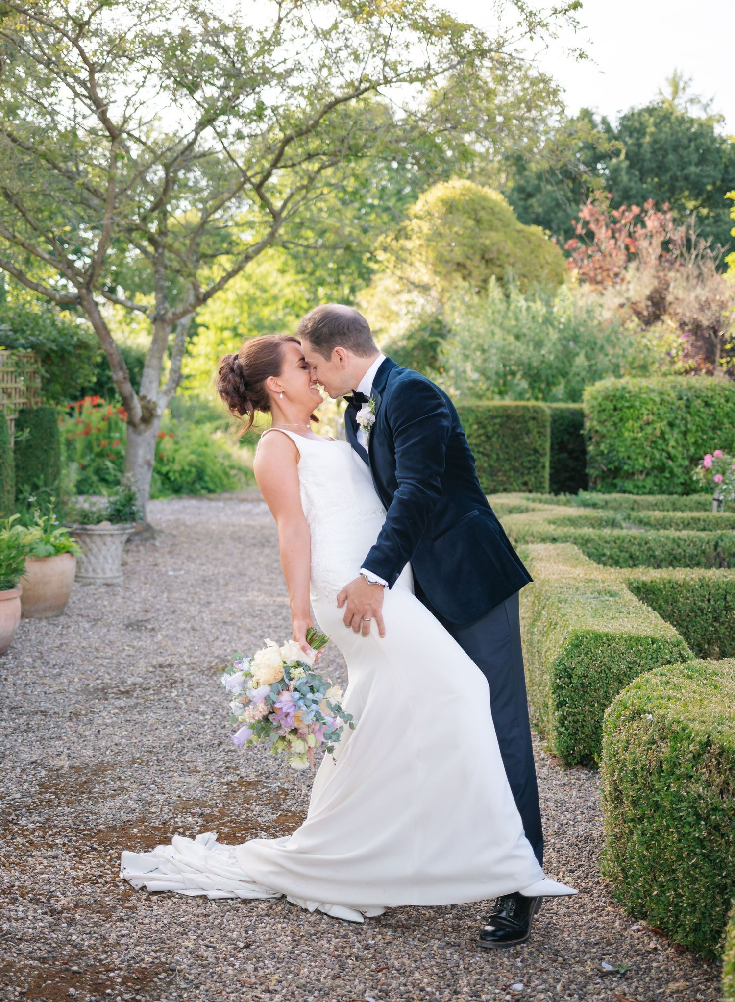 Romantic wedding couple embraces on garden path surrounded by manicured hedges at sunset.