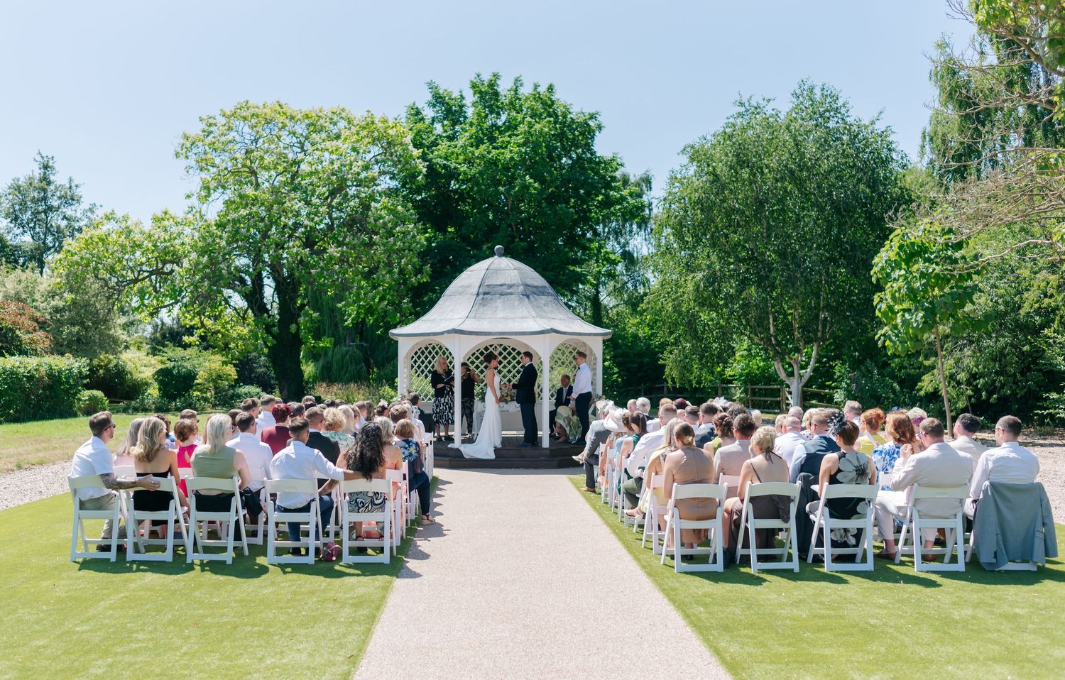 Panoramic view of an outdoor wedding ceremony setup with white chairs and gazebo surrounded by lush green lawn.