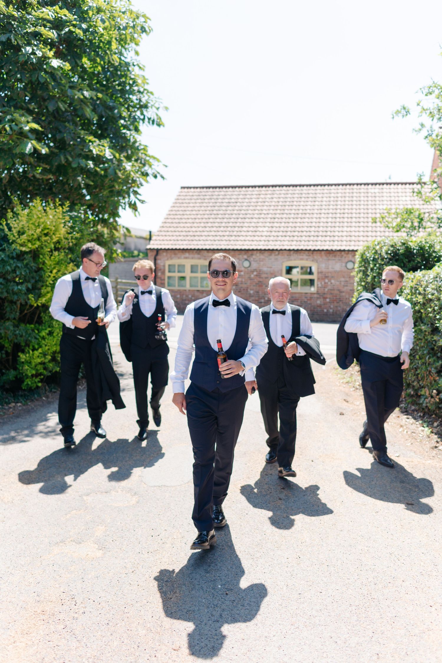 Groomsmen in black vests and bowties perform a playful jumping sequence outdoors.