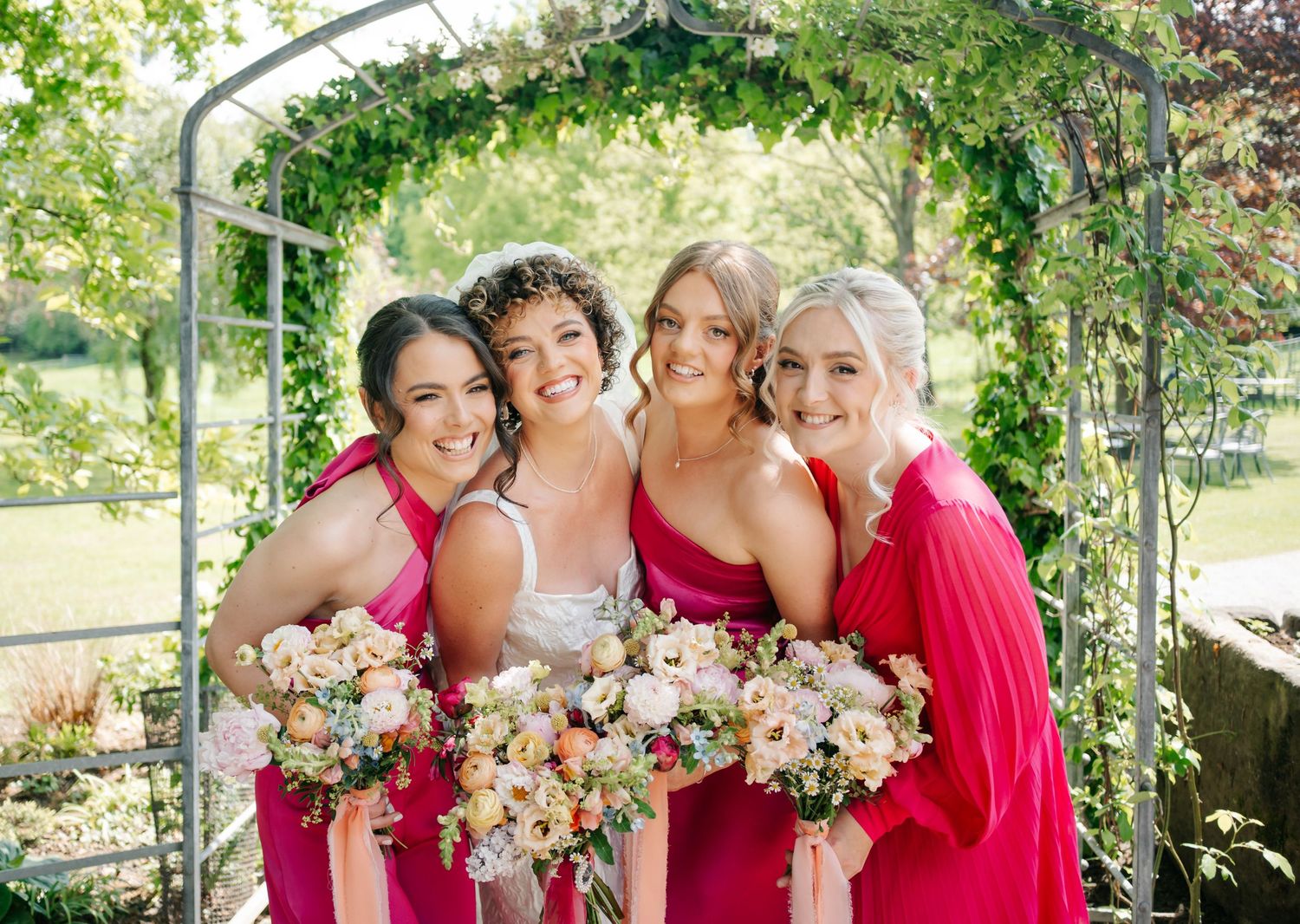 Bridesmaids in bright pink dresses pose under a vine-covered garden arch with bouquets.
