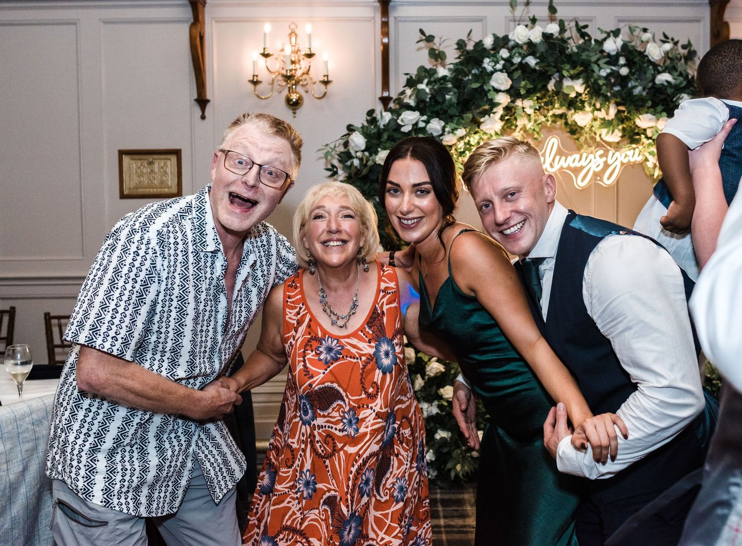Wedding party members posing playfully under neon sign at reception.