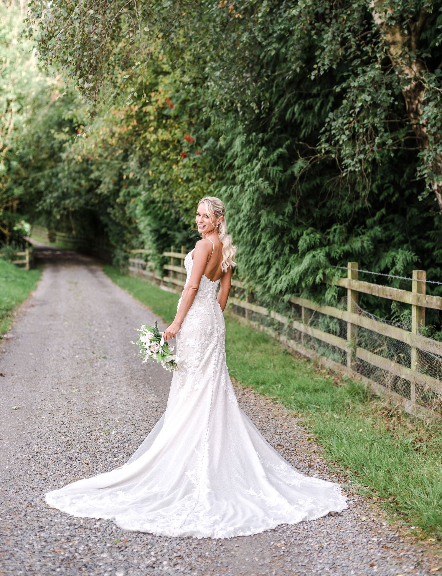 Bride in white lace gown with train poses on country path near wooden fence.