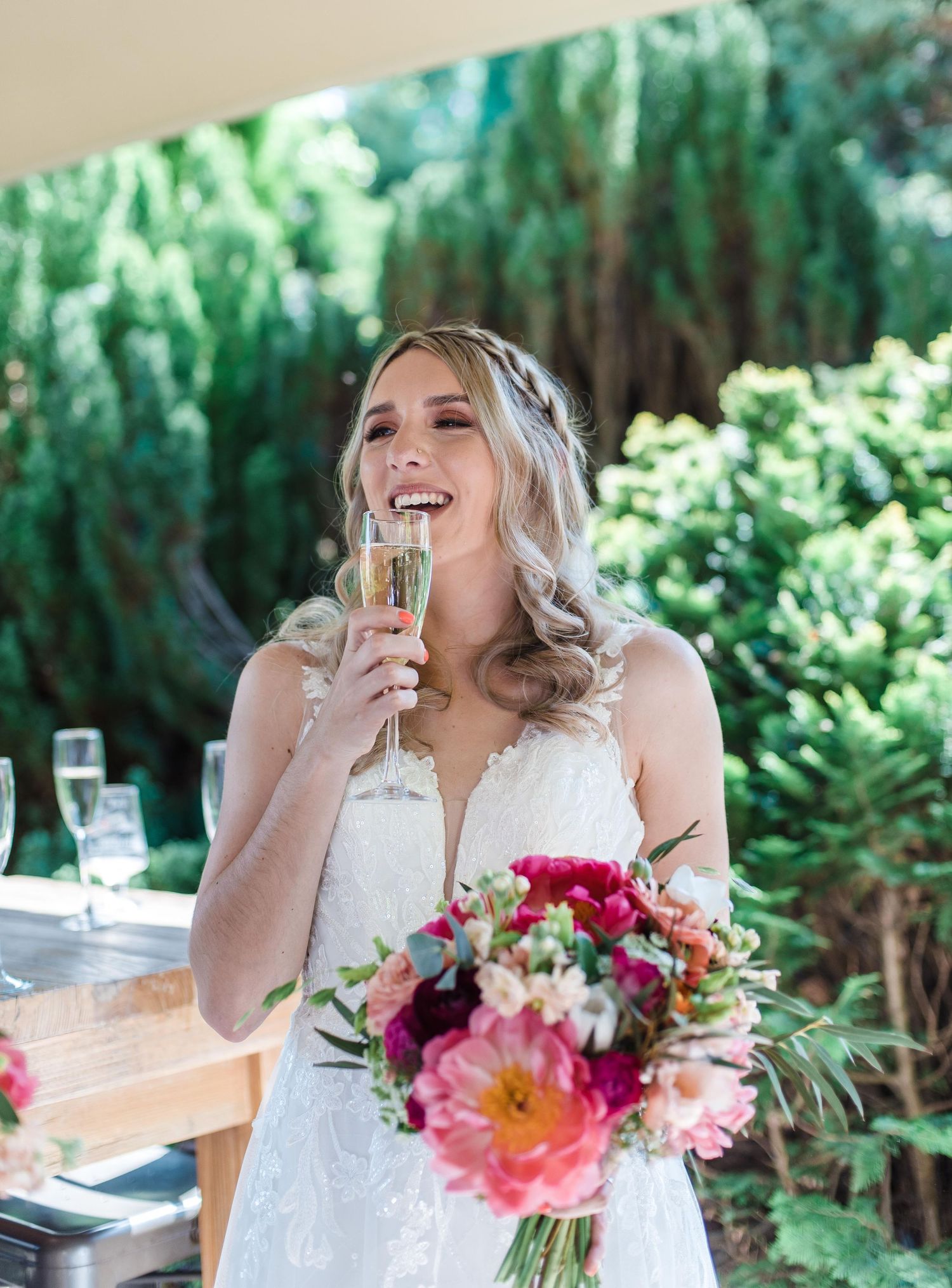 Bride in white dress holding bouquet and champagne glass in garden setting.