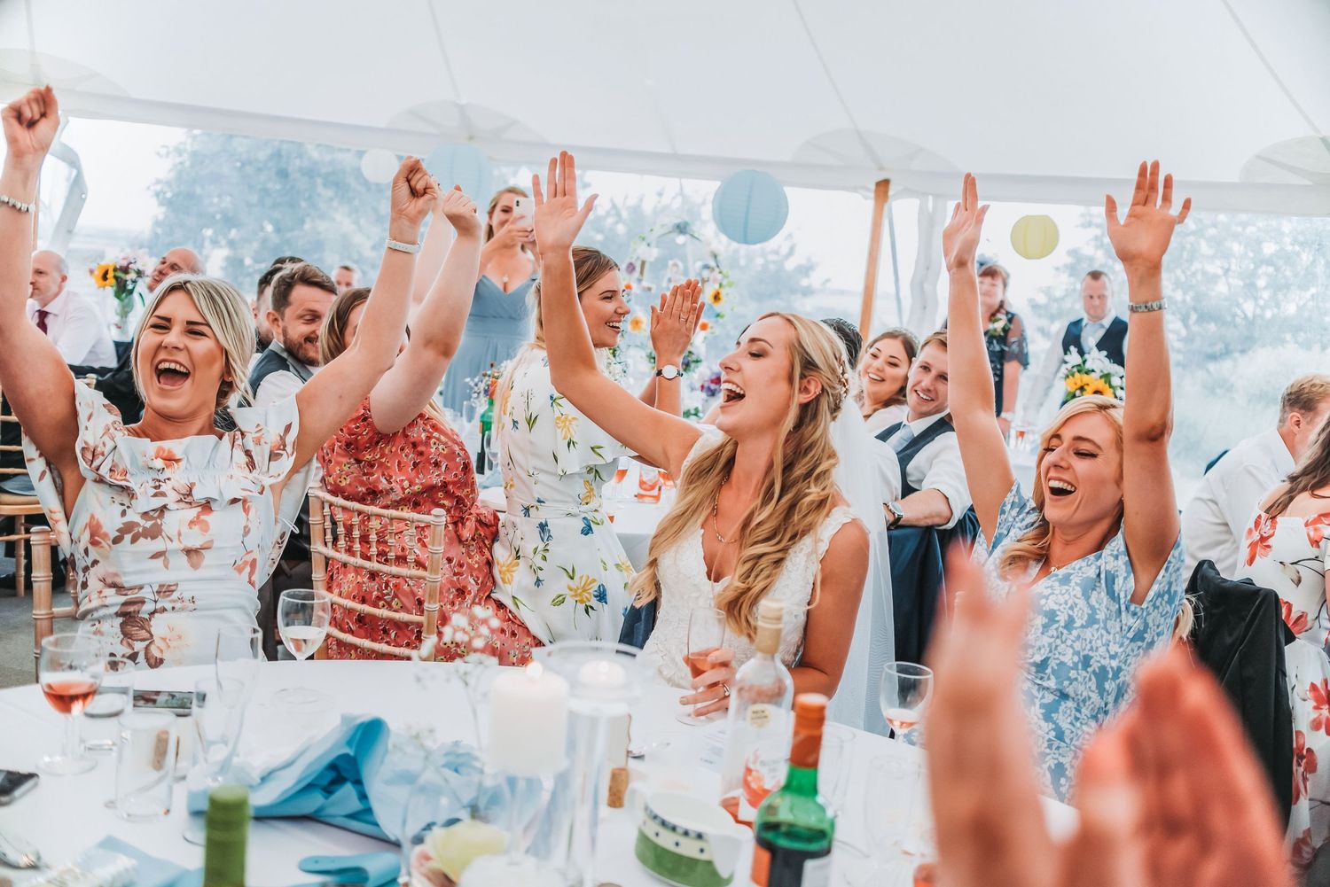 Wedding reception guests cheering with raised arms under white tent with blue lanterns.
