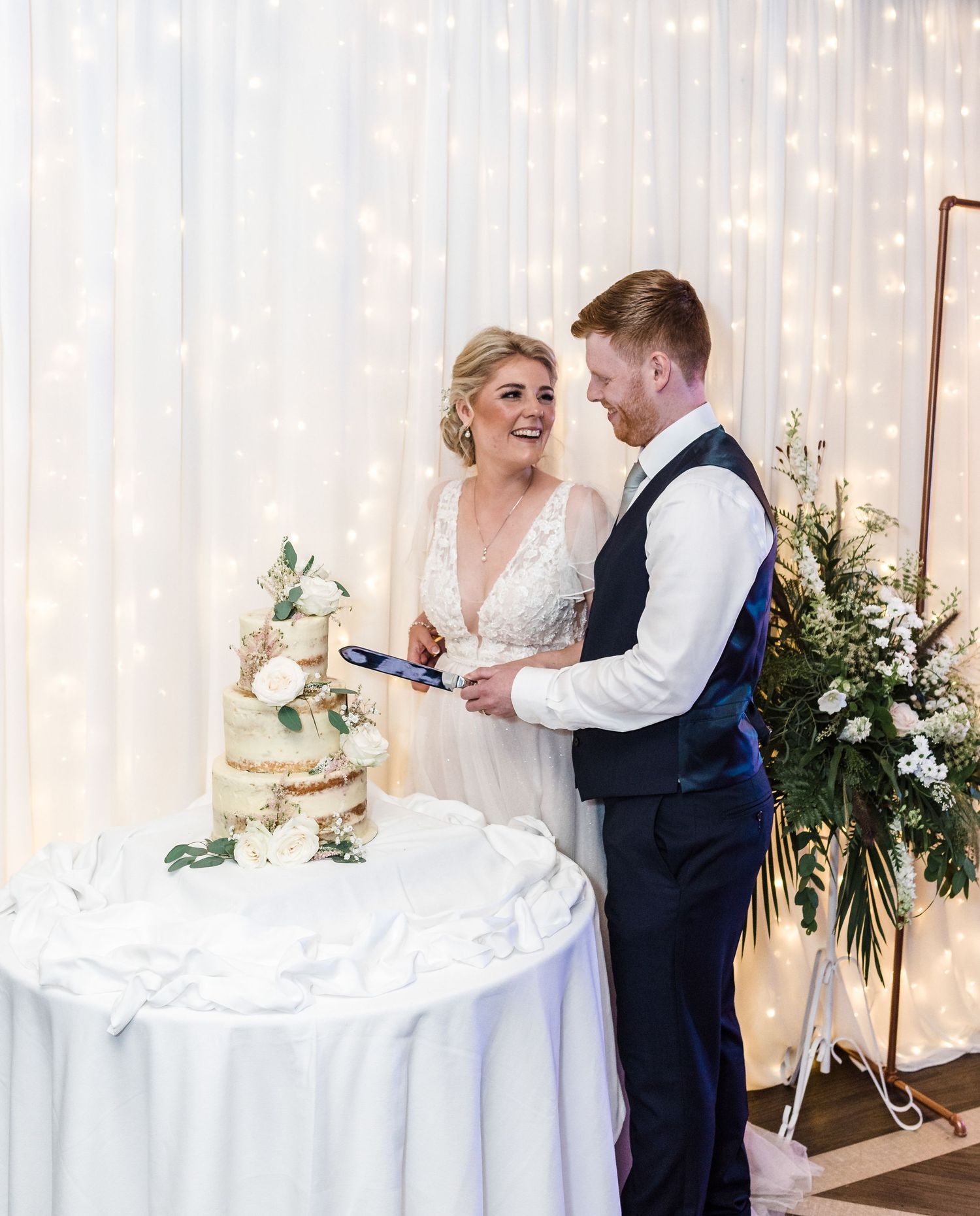 A couple cuts their elegant semi-naked wedding cake together in front of a sparkly backdrop.