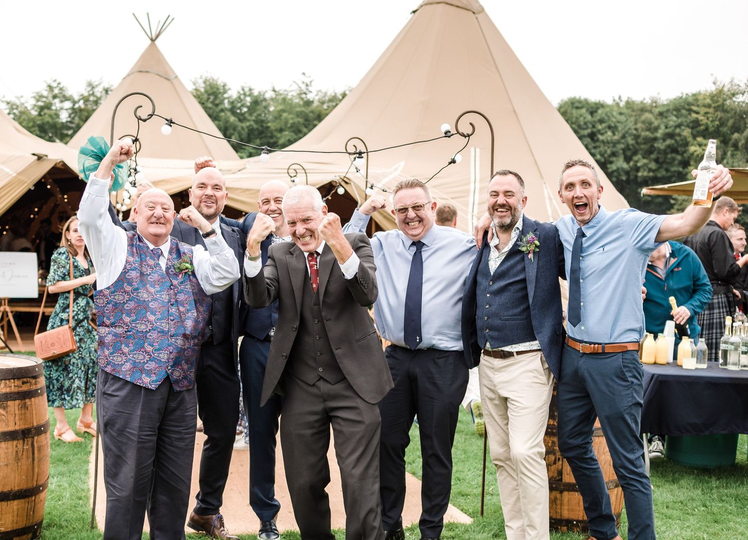 A celebratory group photo at an outdoor wedding reception with a teepee tent in the background.