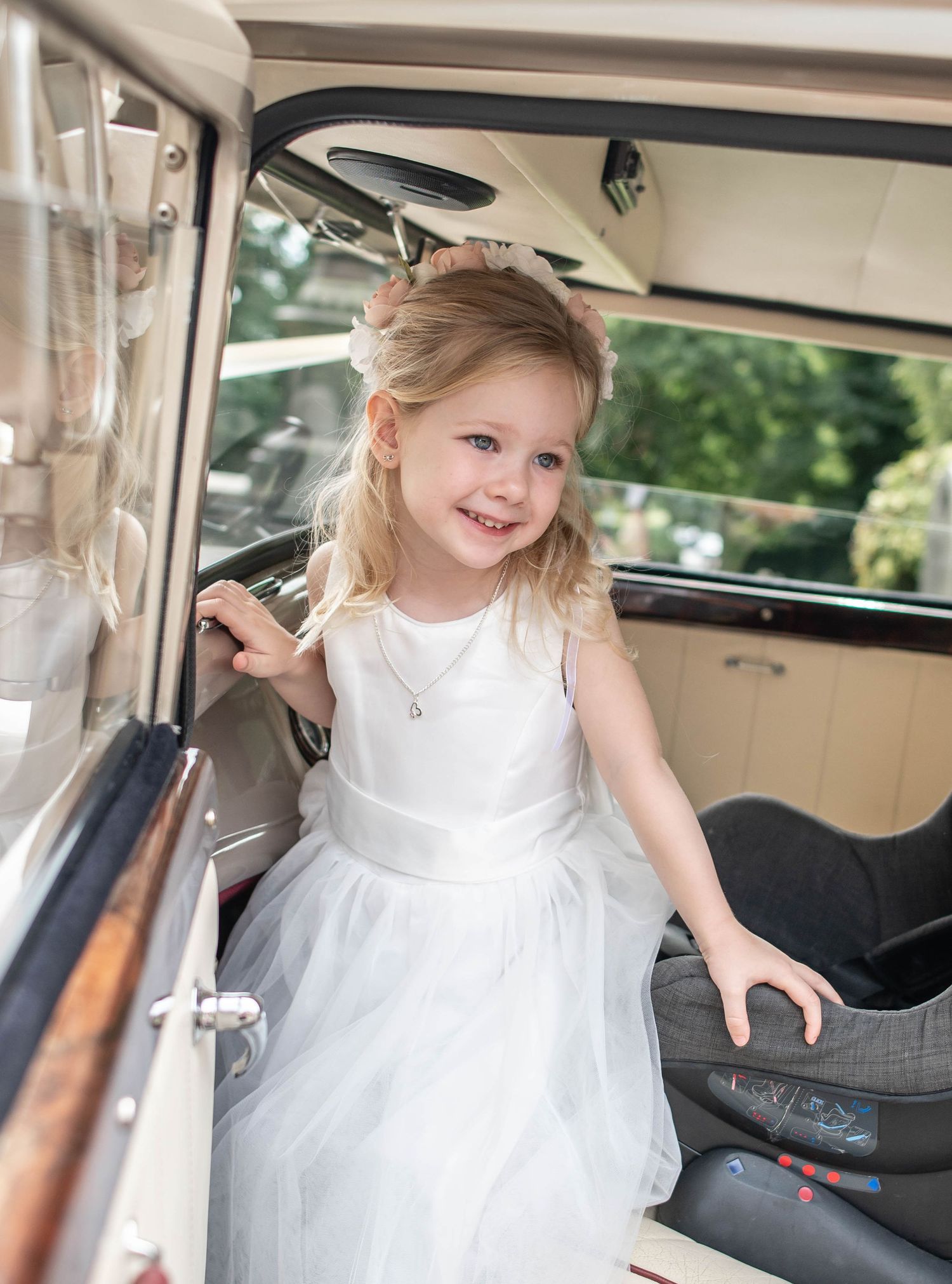A flower girl in a white dress smiles while getting into a classic vintage car.