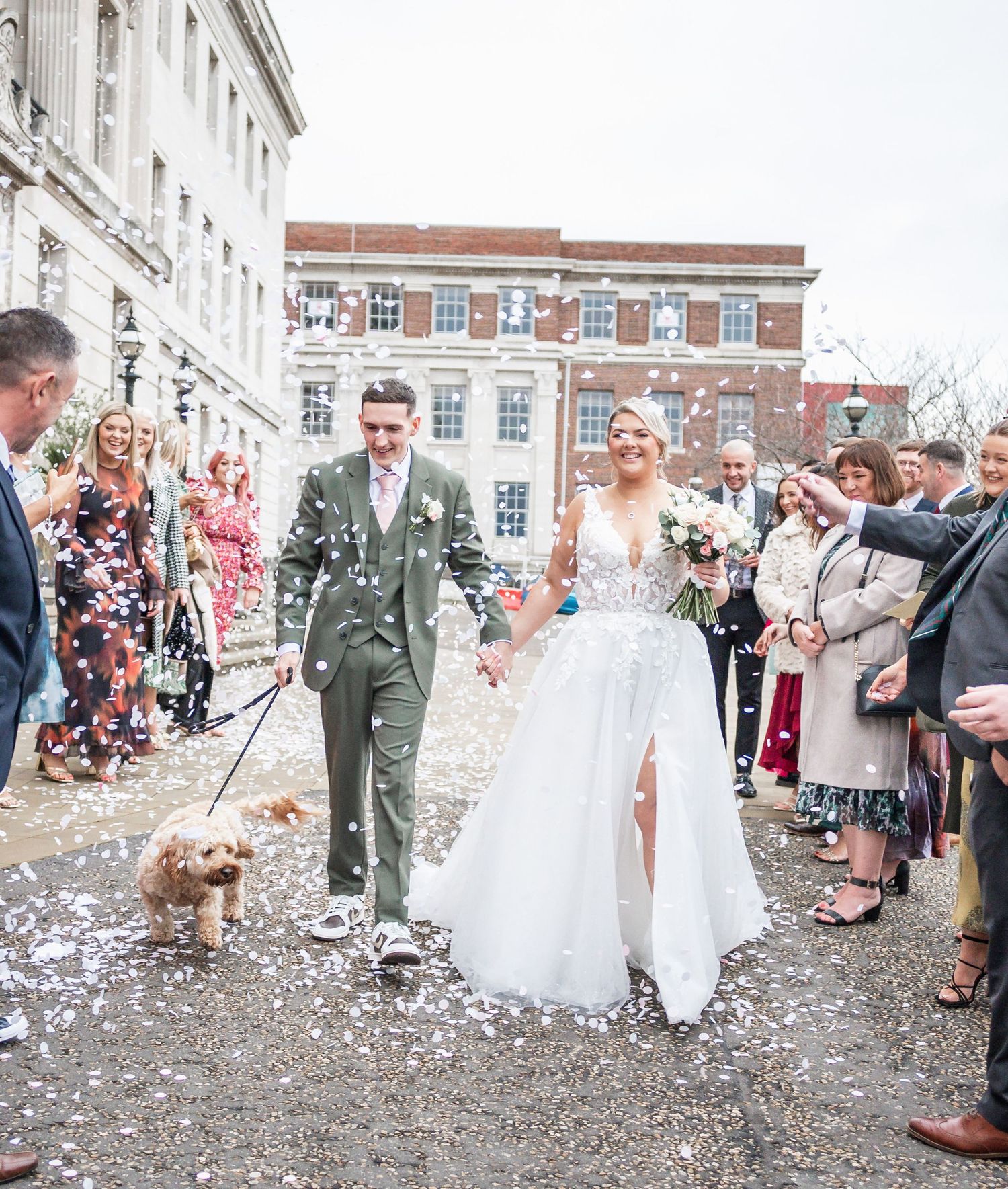 Wedding guests throw confetti as couple walks with their dog through celebratory crowd.