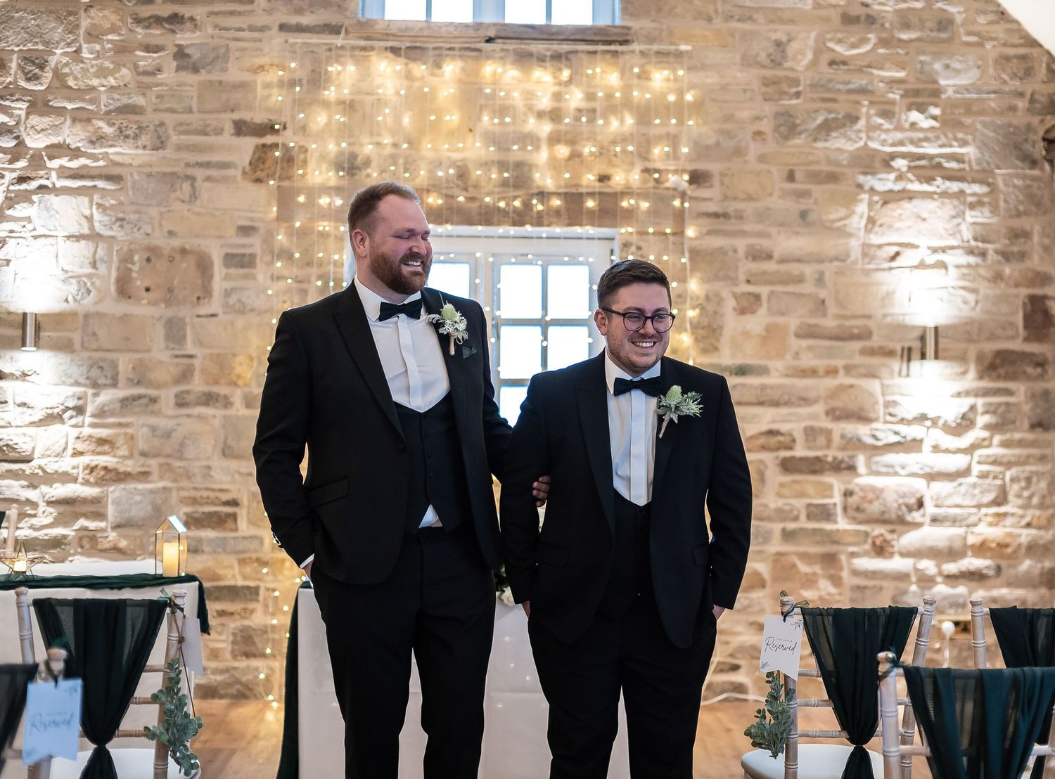 Two people in formal black tuxedos standing against rustic stone wall with string lights.