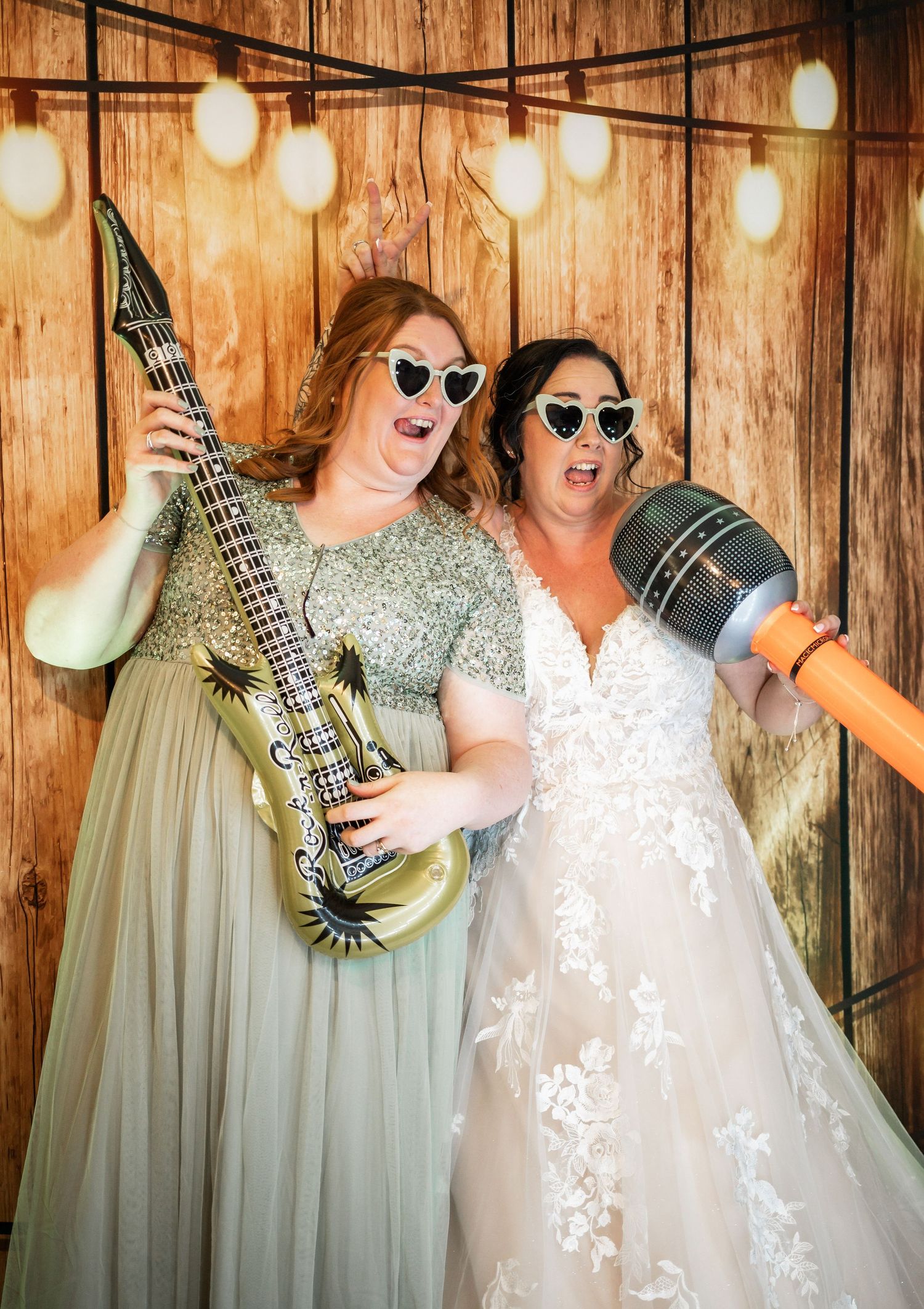 Two wedding guests pose with a guitar and microphone against a rustic wood wall with string lights.