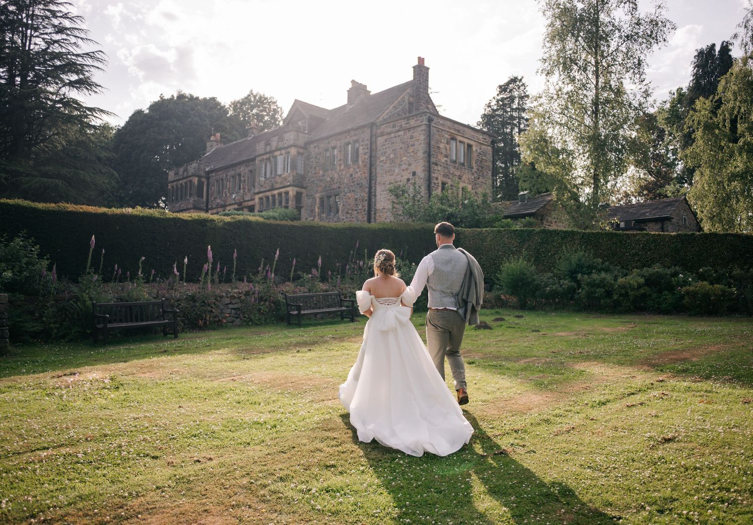 Newlywed couple walks across sunlit lawn with historic stone manor house in background.