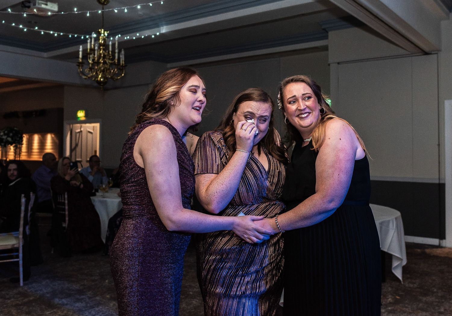 Three wedding guests sharing emotional moment on dance floor under chandelier lighting.