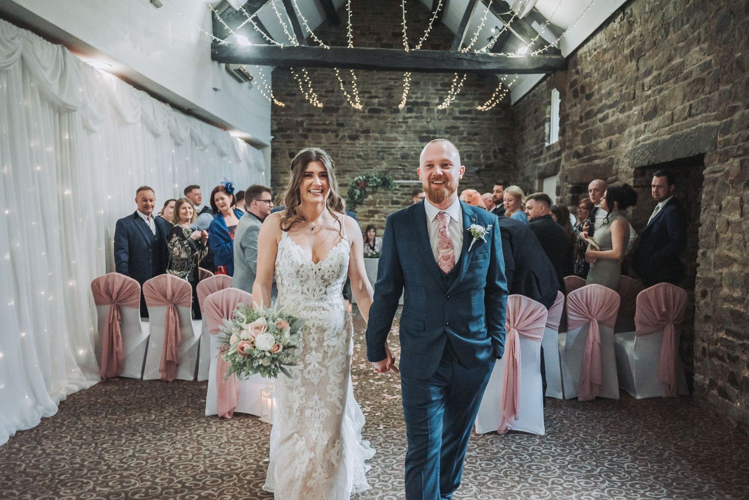 Wedding couple walks down aisle in rustic venue with exposed brick and pink chairs.