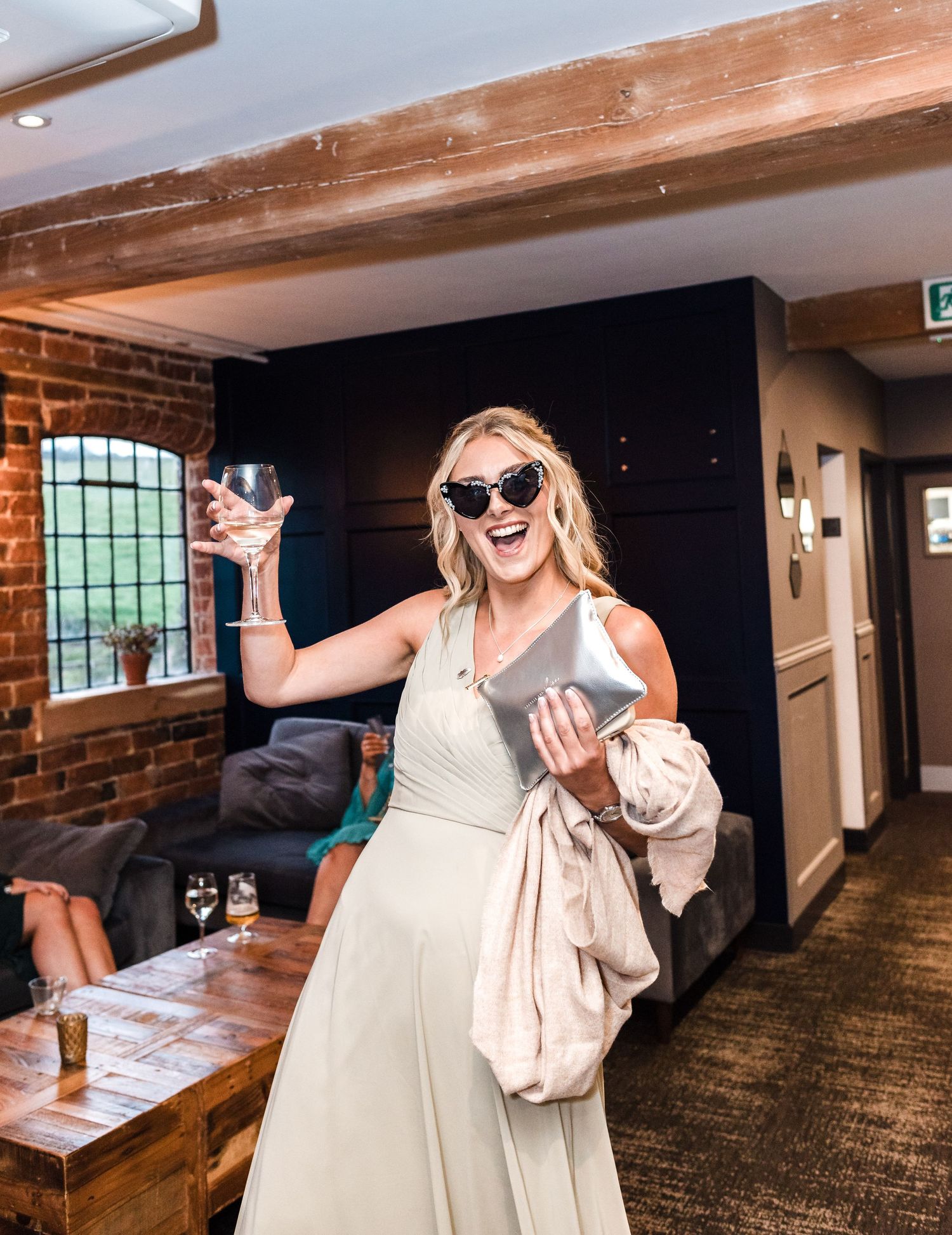 Wedding guest in sunglasses raises glass while celebrating in rustic venue.
