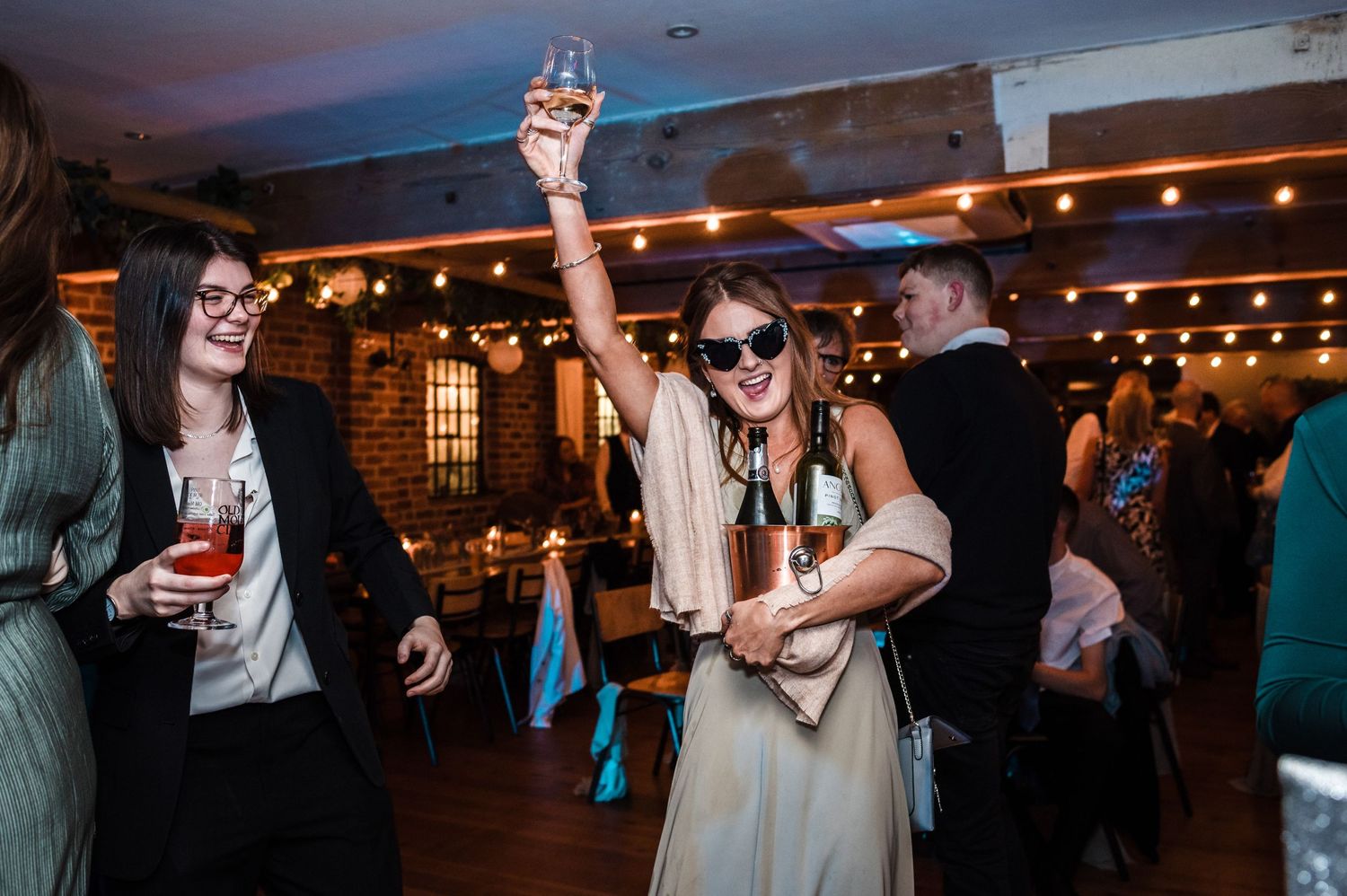 Wedding guests dancing and celebrating with drinks at rustic brick venue with string lights.