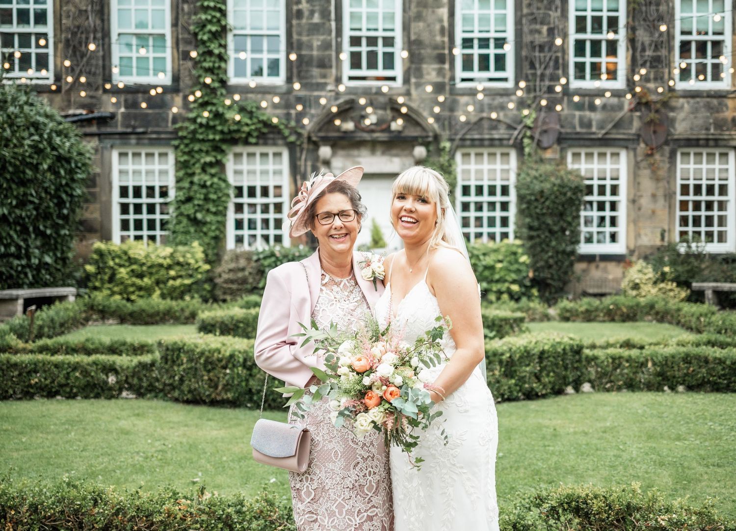 Wedding portrait in front of ivy-covered building with manicured gardens.