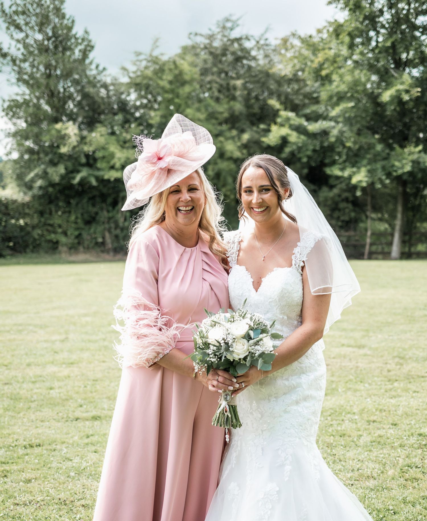 A bride and guest in pink enjoy a sunny moment in a garden setting.