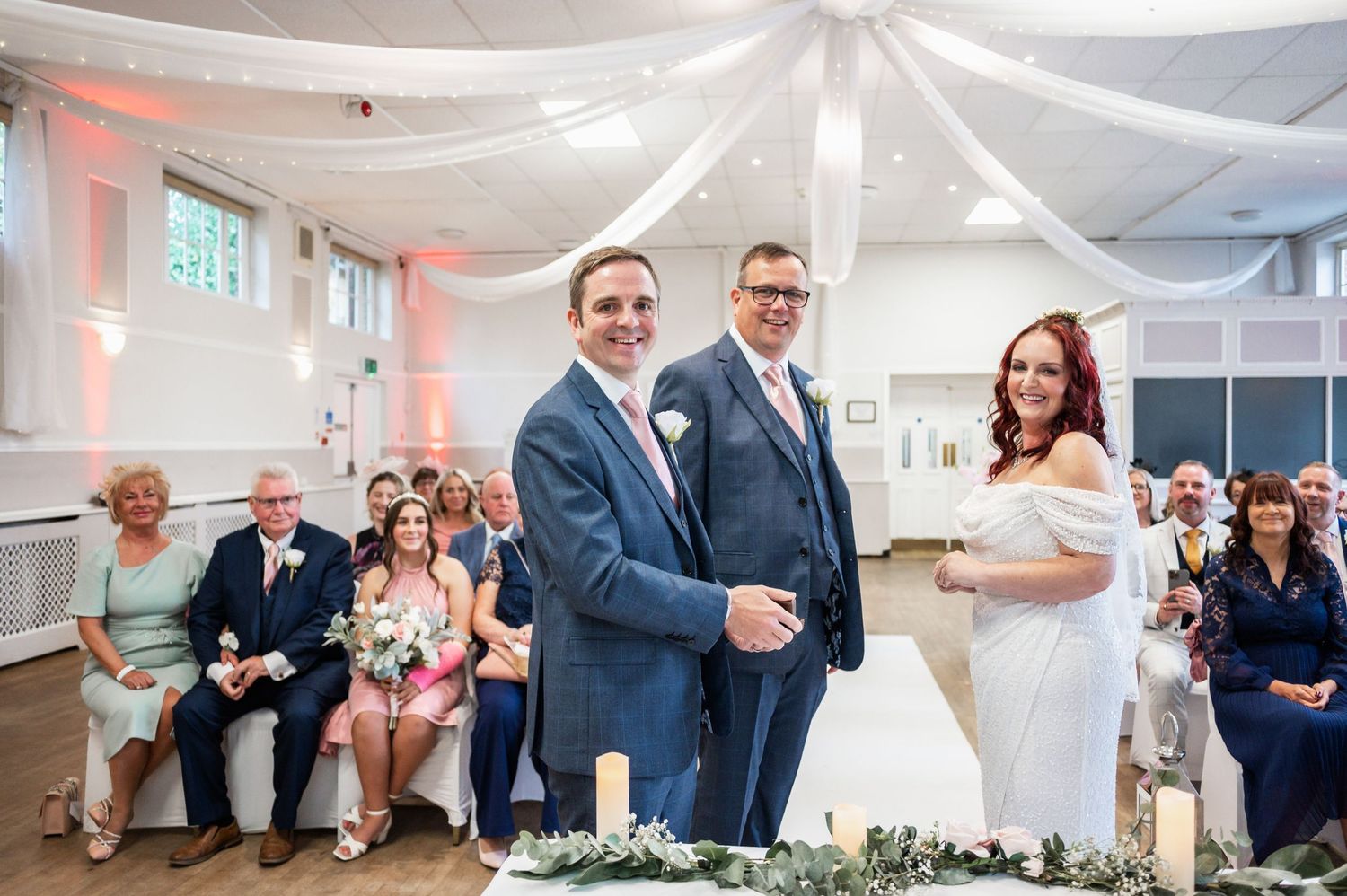 Wedding ceremony in a white decorated room with pink accents and gathered guests.