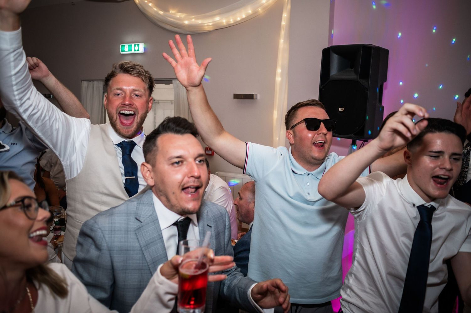 Enthusiastic wedding guests dancing and raising their hands at a lively reception party.