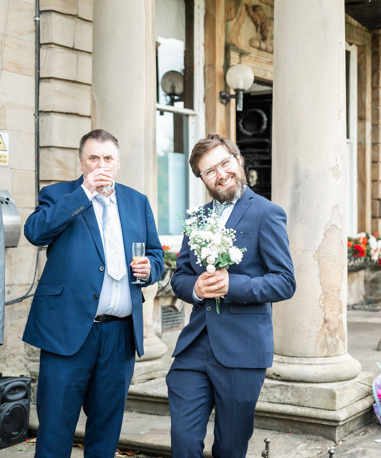 Wedding guests in navy suits sharing drinks outside venue with classical columns.