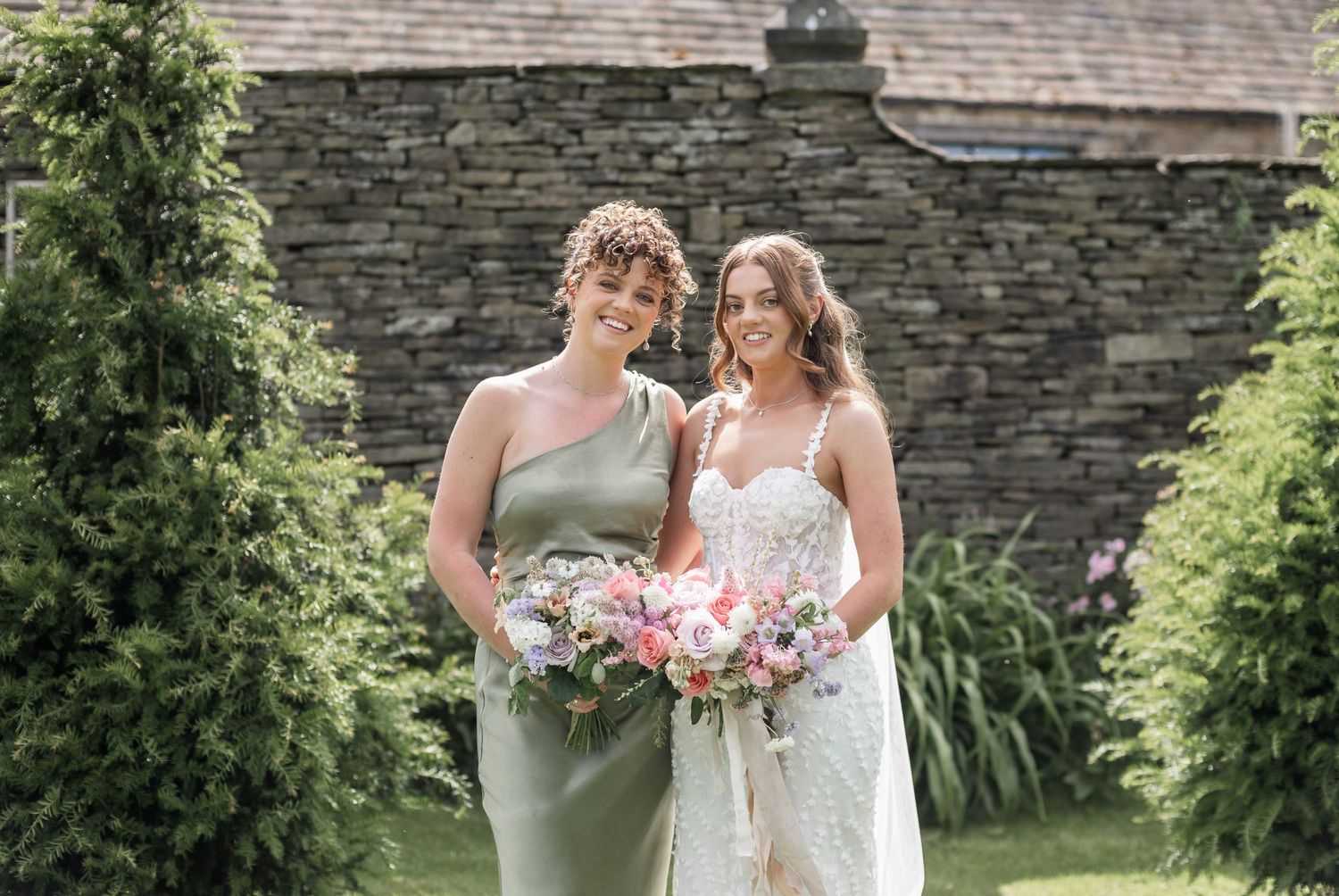 Two attendants pose by stone wall with lush floral arrangements in garden setting.